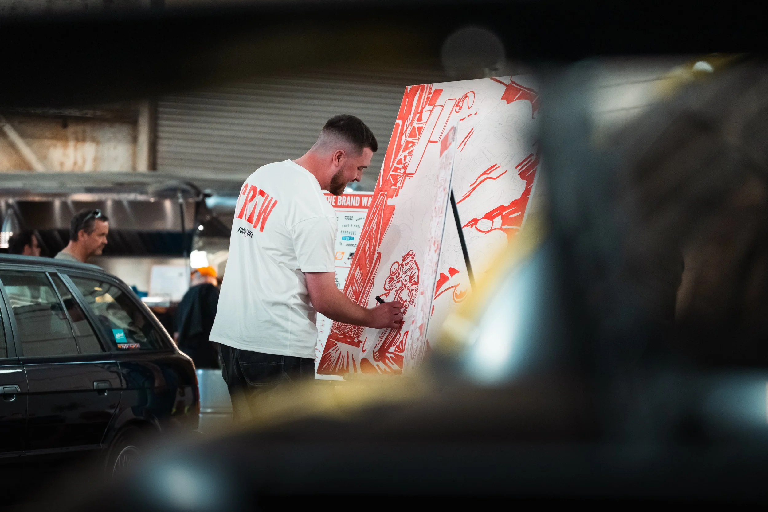 A man signing a large poster with a red and white design at an indoor event. There are other people and cars visible in the background.