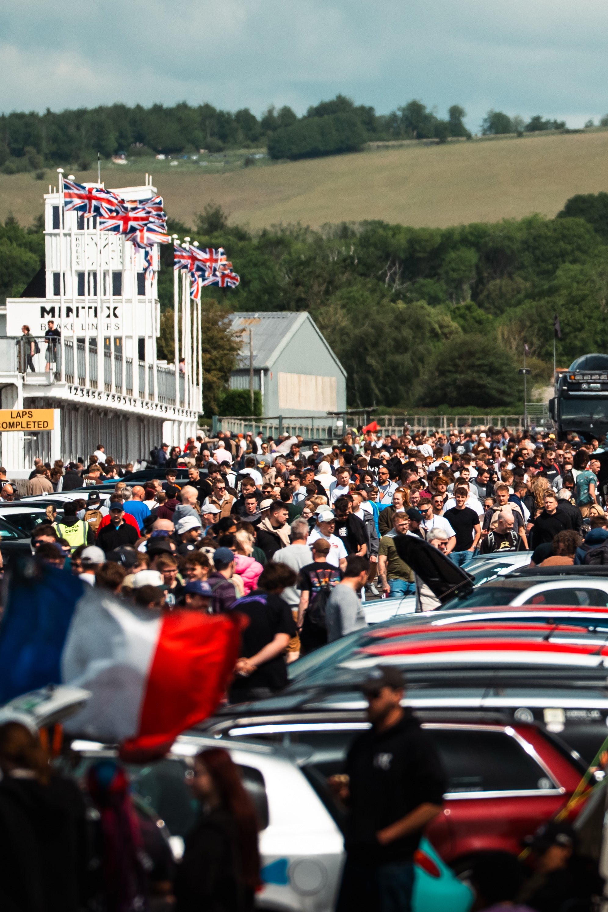A crowded parking lot at a racing event with many cars and people. In the background, there is a building with a sign that says 'MINTEX' and a row of Union Jack flags.