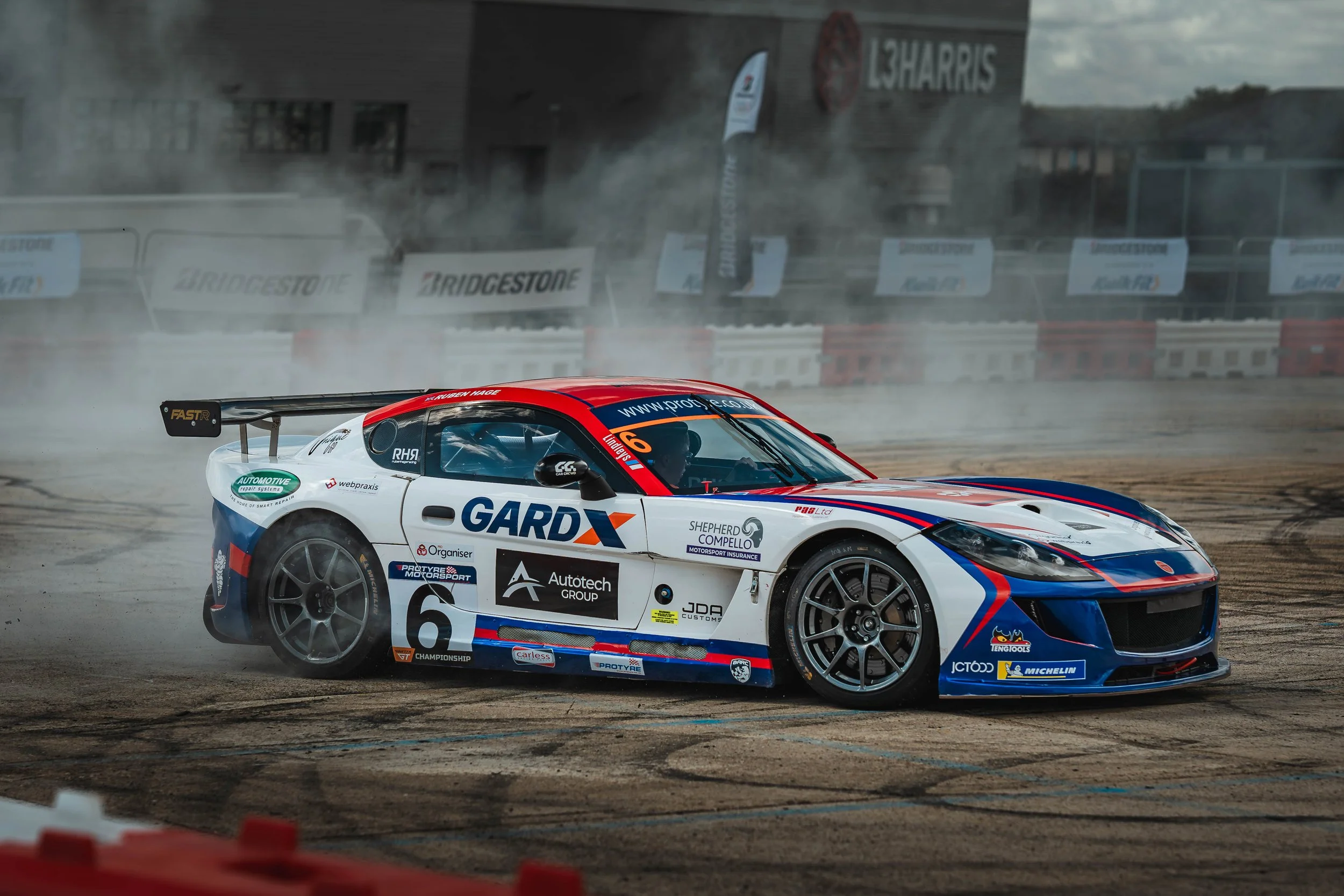 Race car drifting on track with smoke, sponsor logos, and a building with '13HARRIS' sign in background.