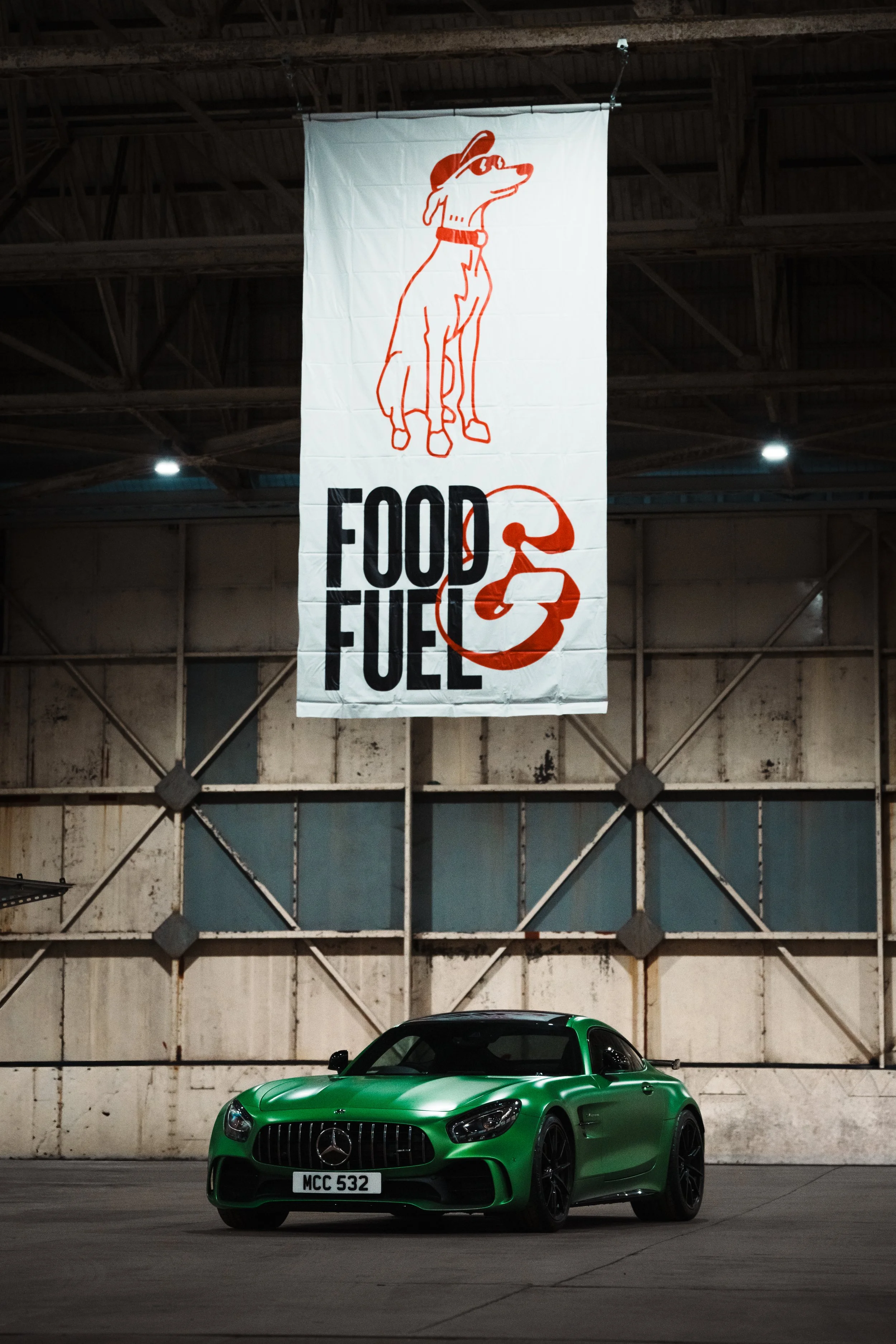 A green Mercedes-Benz sports car with a custom license plate 'MCC 532' parked in front of a large white banner with an illustration of a dog wearing sunglasses and a hat, along with the words 'FOOD & FUEL' displayed below the dog.