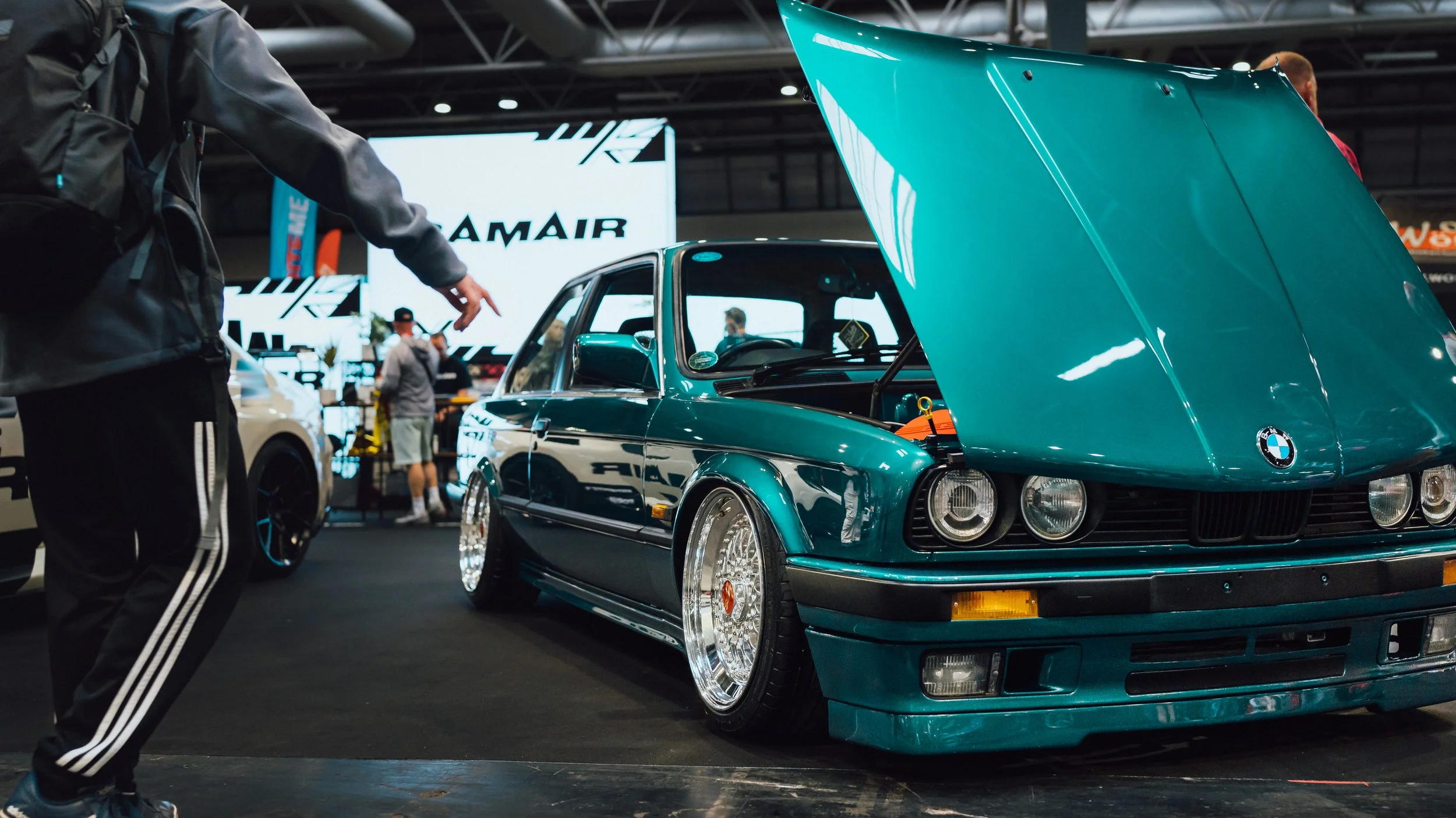 A teal vintage BMW car with the hood open at an indoor car show.