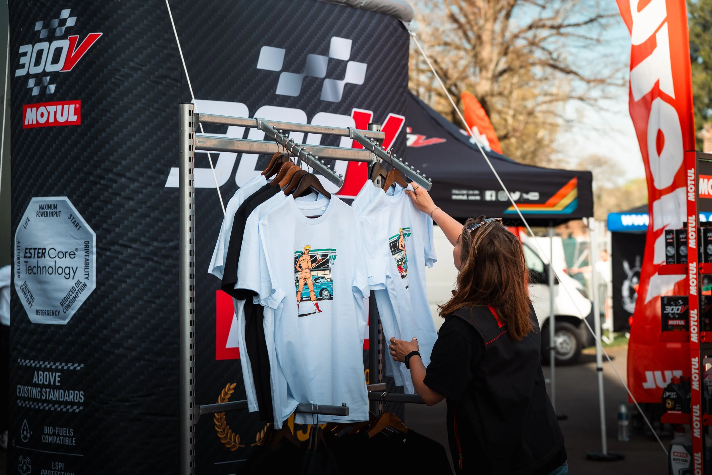 A woman with sunglasses on her head shops for graphic T-shirts at an outdoor event. The T-shirts feature a cartoon of a woman in a uniform, standing next to cars with a city street background. The scene is set under a black tent banner with racing an