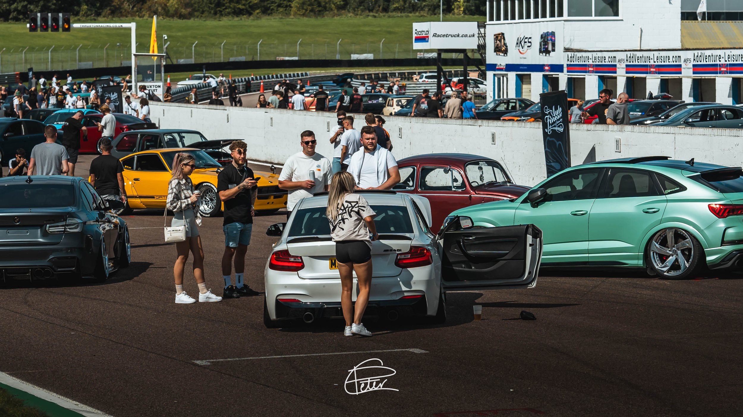 People at a car meet at a race track, with various cars parked and spectators socializing.