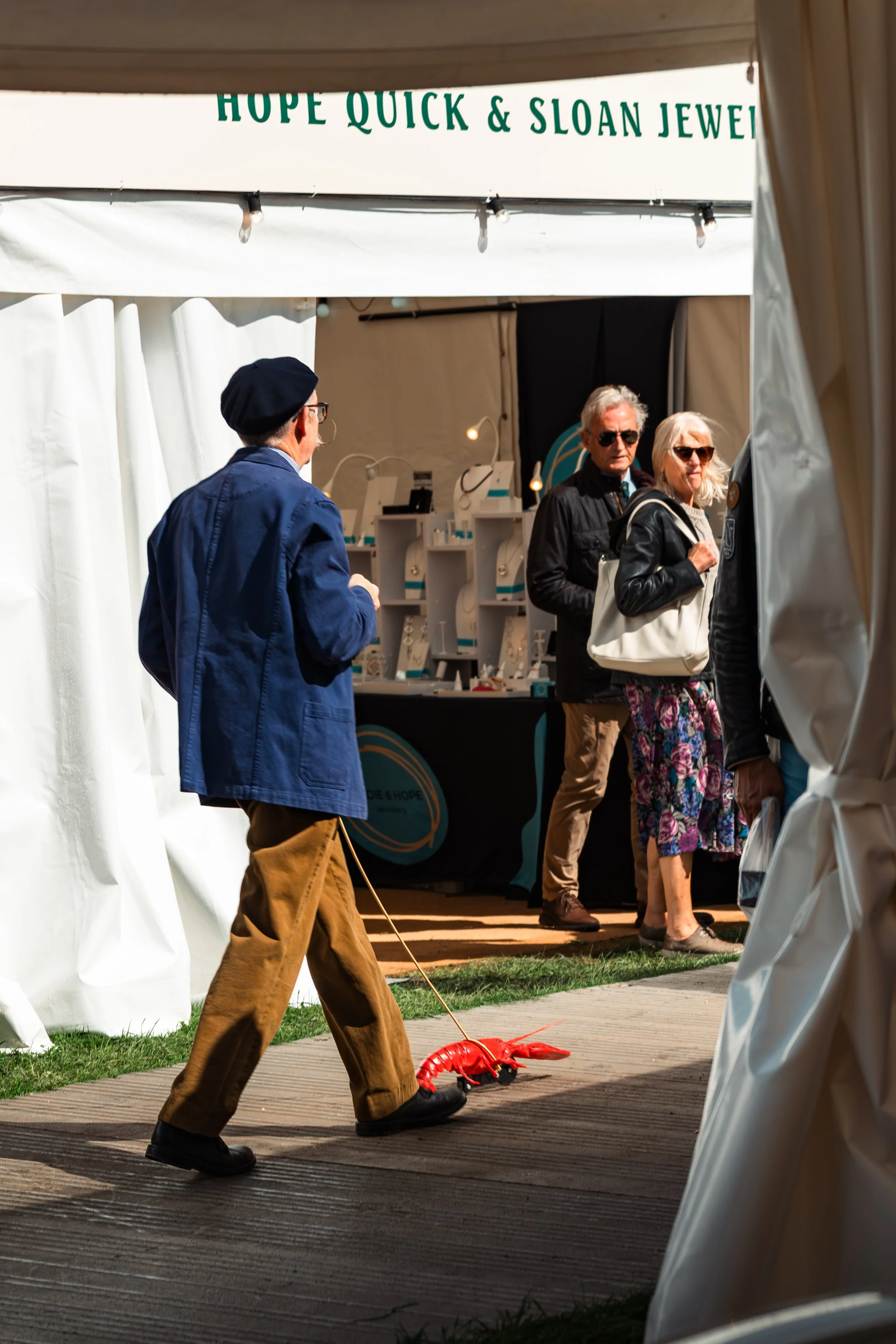 People line up at a jewelry booth under a tent with a sign that reads 'Hope Quick & Sloan Jewelry.' A man in a blue jacket and tan pants walks past a red lobster-shaped pulling toy on the ground, while other people browse inside the booth.