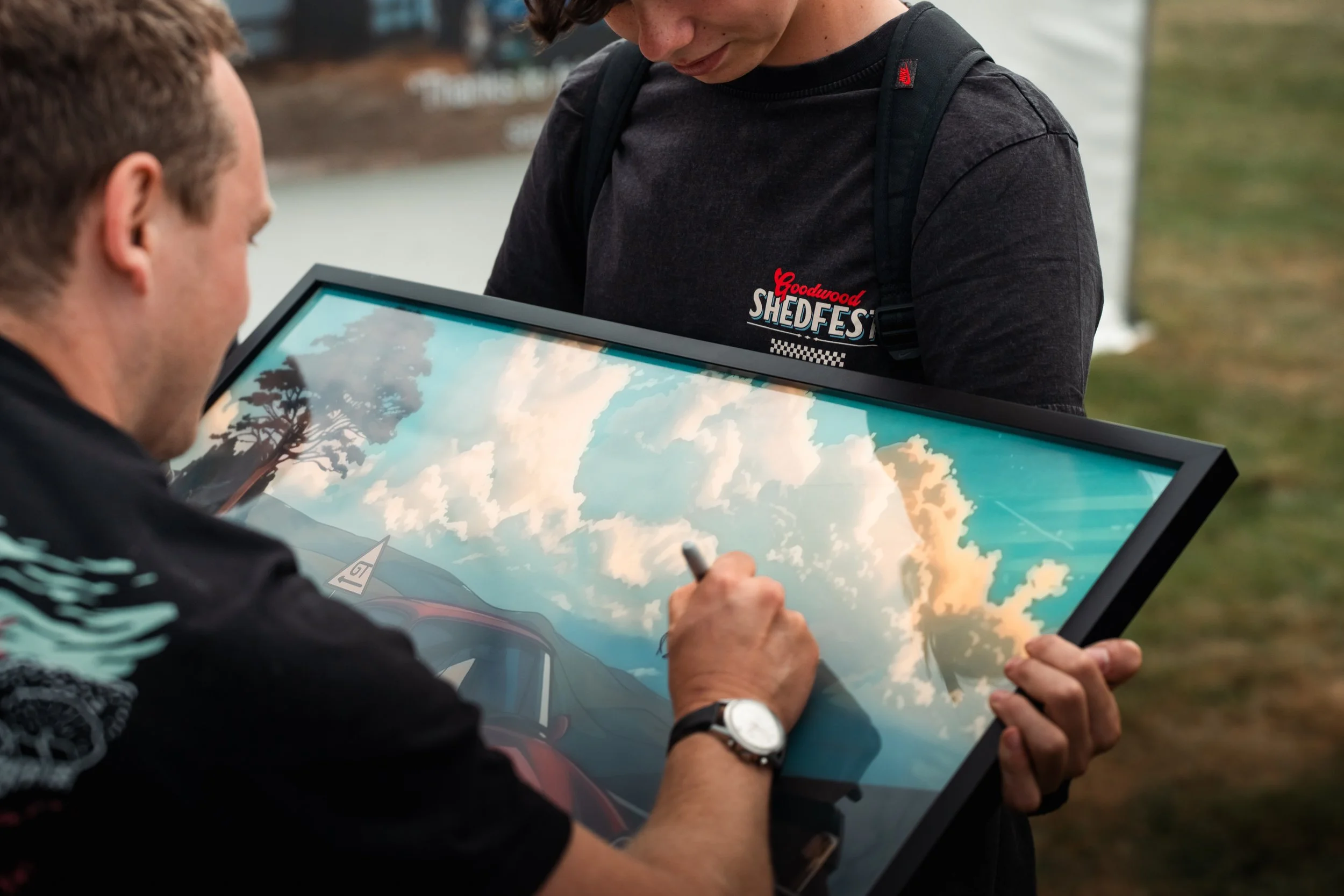 Two men are outdoors, one signing or drawing on a framed picture of sky and clouds, while the other observes.