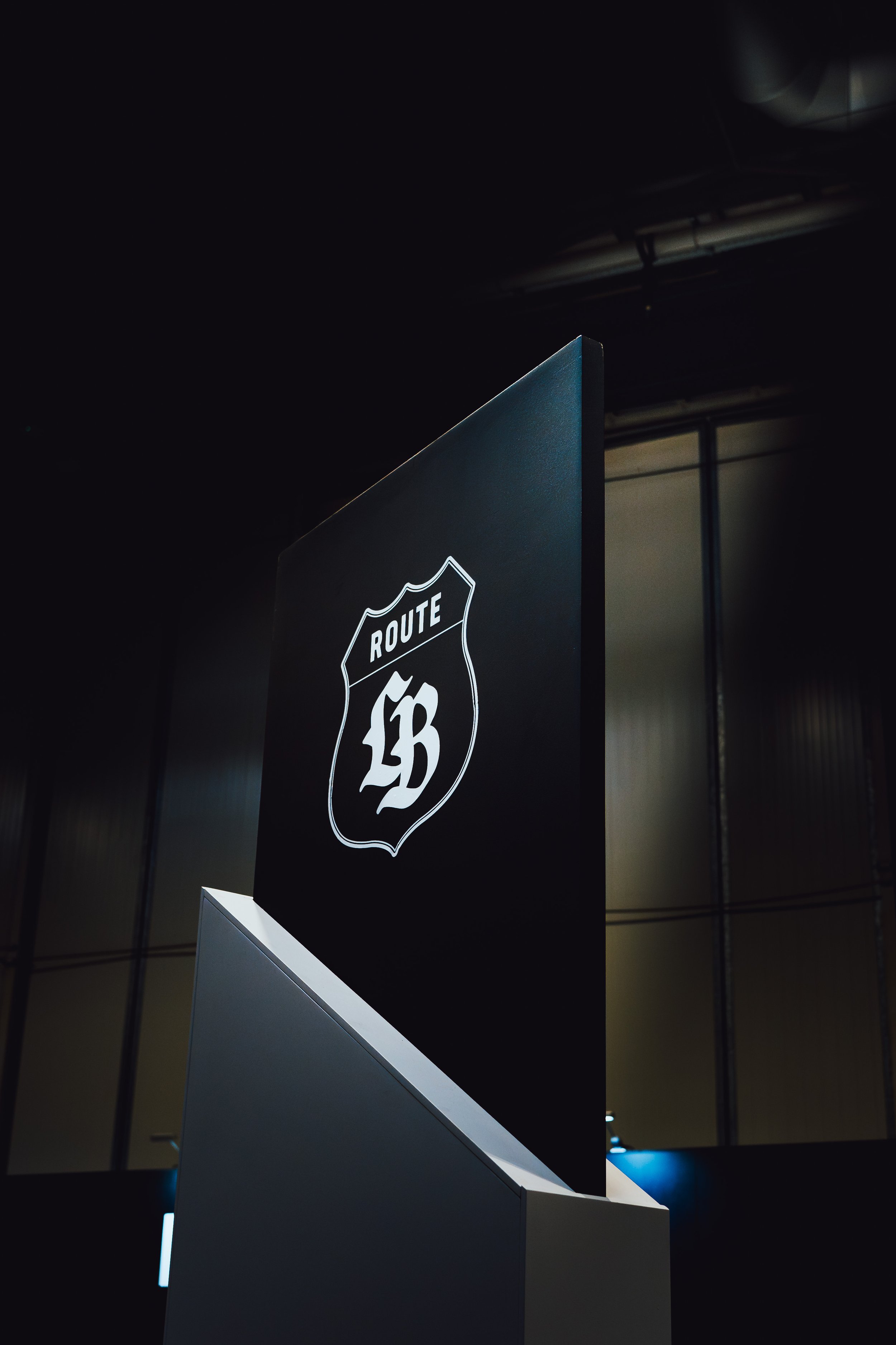 Photo of a black sign with a white Route 66 shield logo, placed on a white base, indoors with visible ceiling and window in the background.