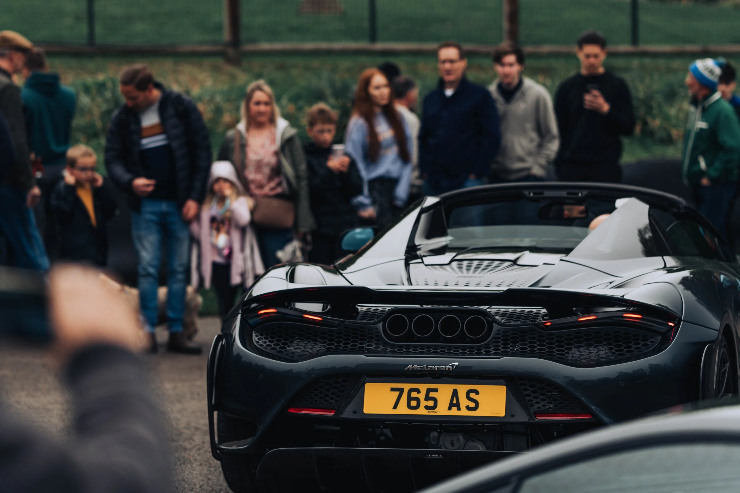 A group of people gathered outdoors, with a black sports car in the foreground.