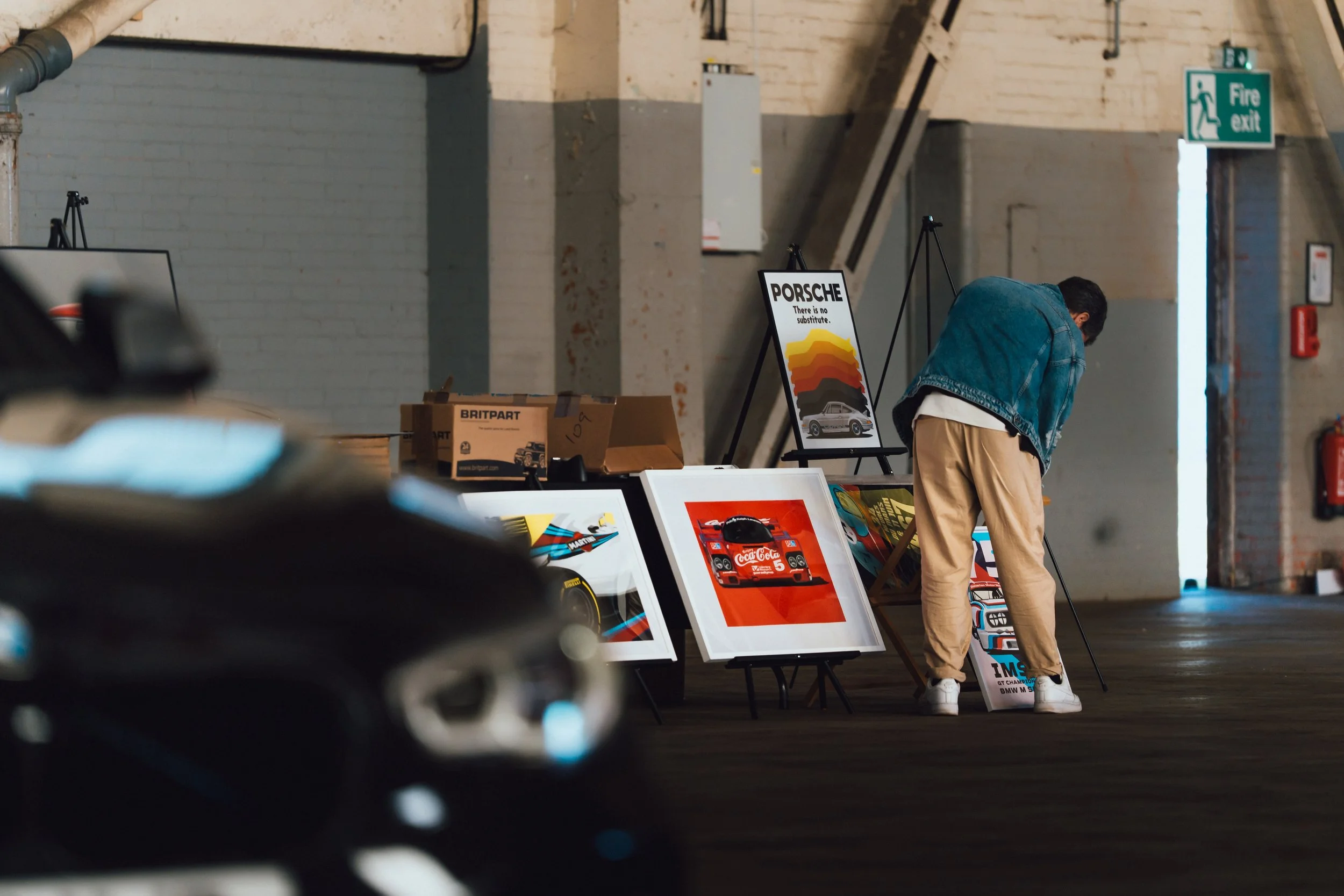 A person bending over and looking at framed posters or artwork on display at an indoor art exhibition, with a Porsche poster and other automotive-themed posters visible.