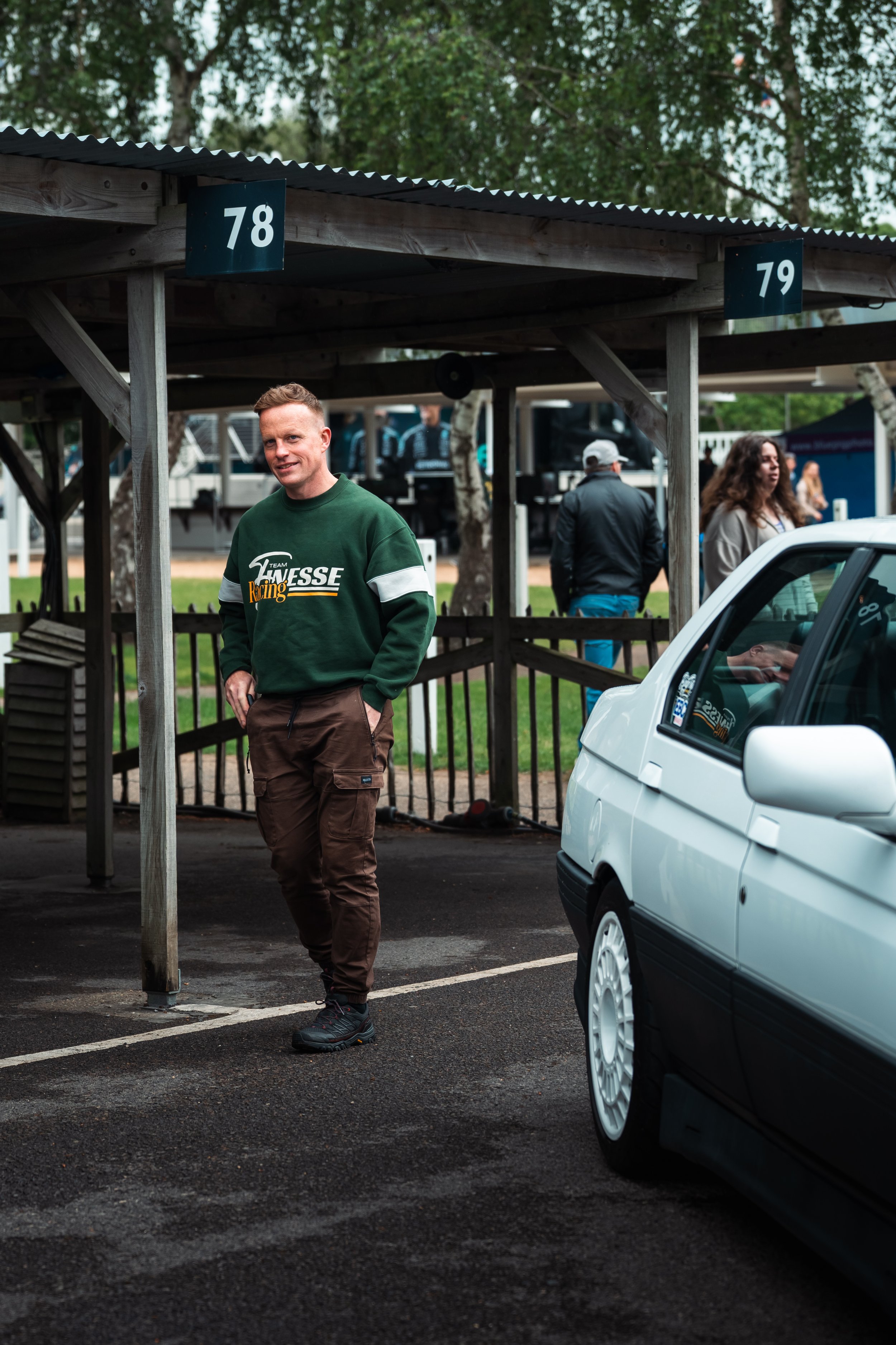 A man dressed in a green racing jacket with brown pants standing in a parking lot under a wooden shelter with numbered signs, next to a white car and other people in the background, with trees and cloudy sky overhead.