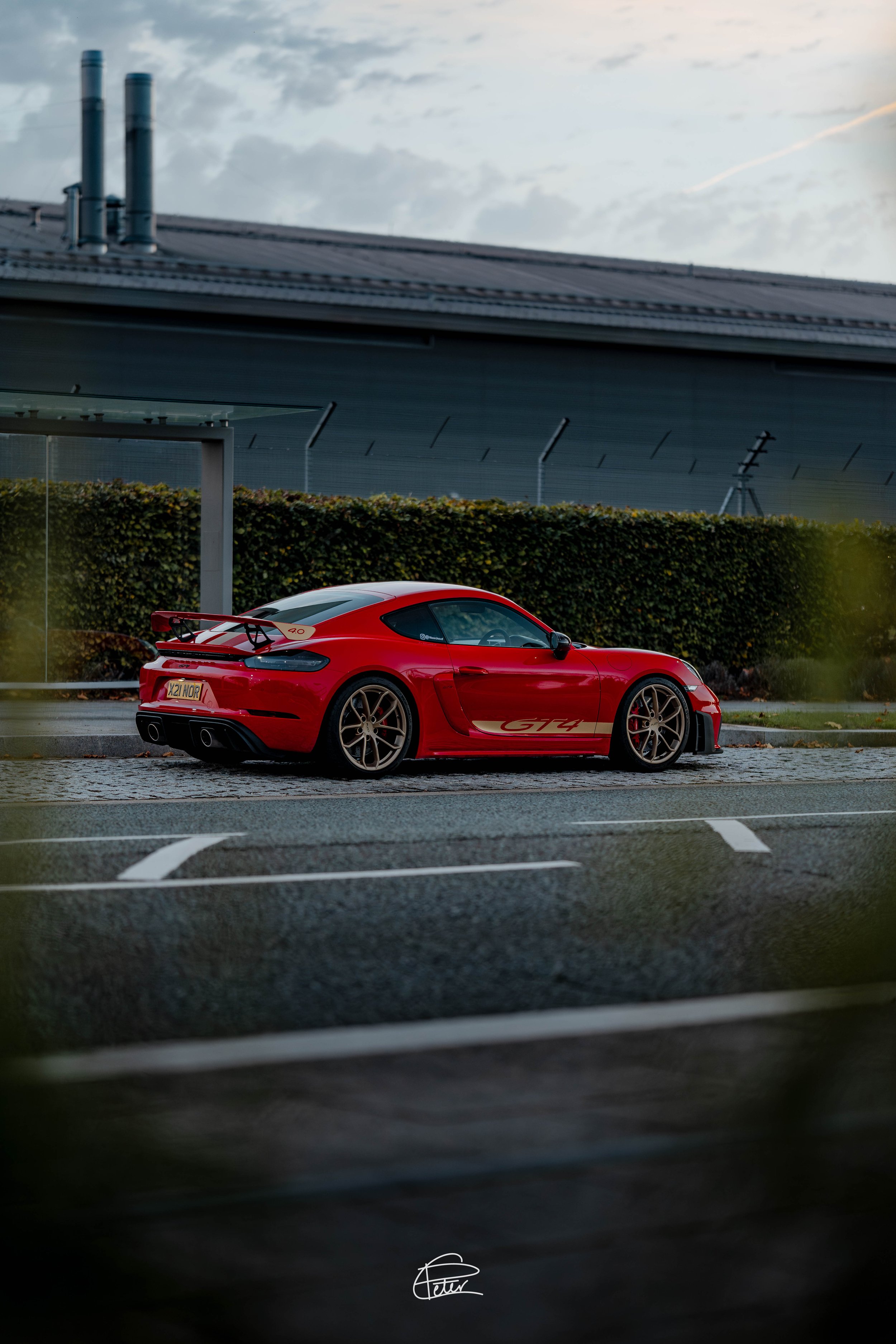 A red Porsche GT4 sports car parked on a street near a green hedge, with a modern building and cloudy sky in the background.