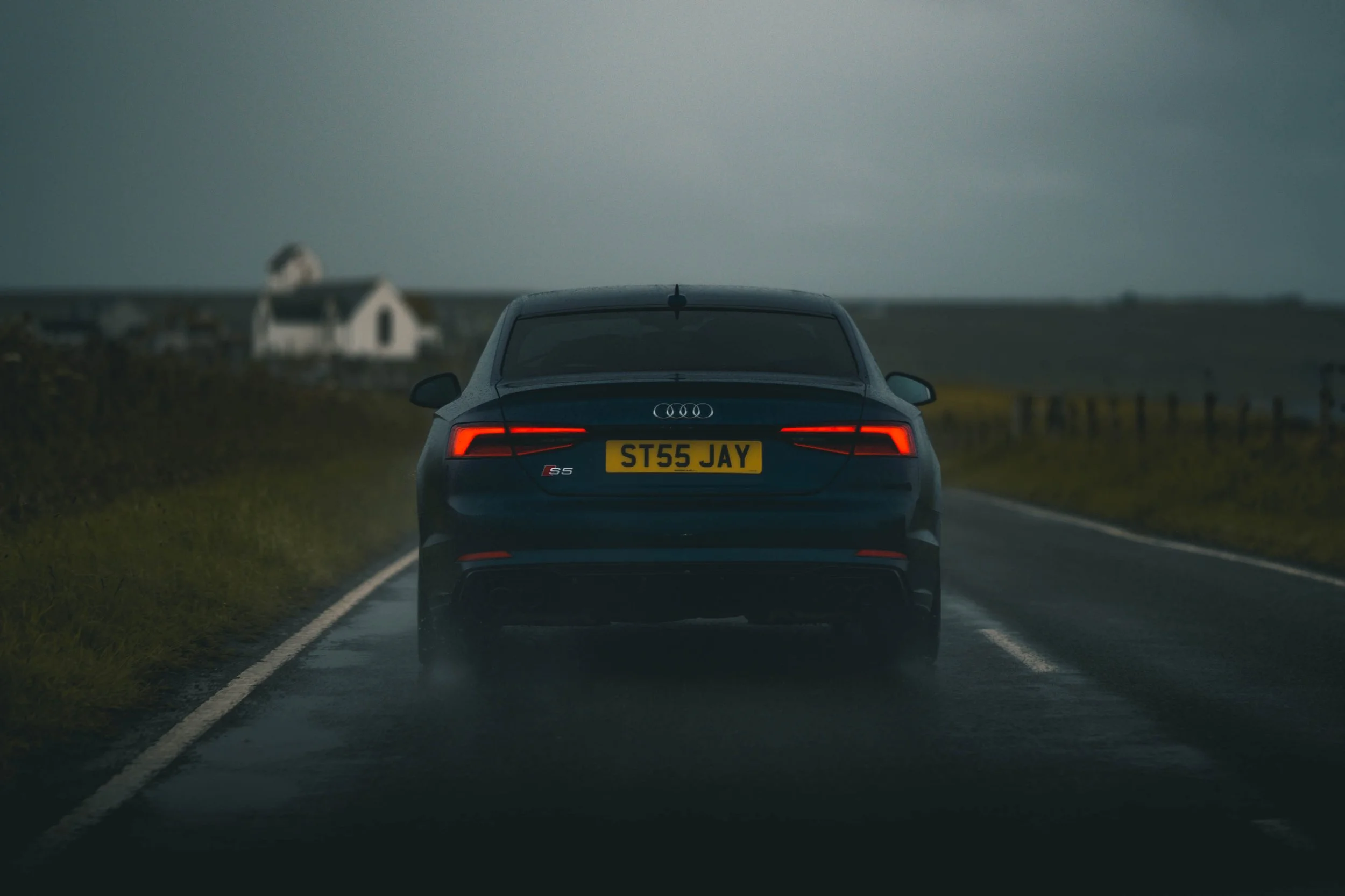 Back view of a black Audi S5 driving on a wet countryside road during dusk or dawn, with blurred fields and houses in the background.