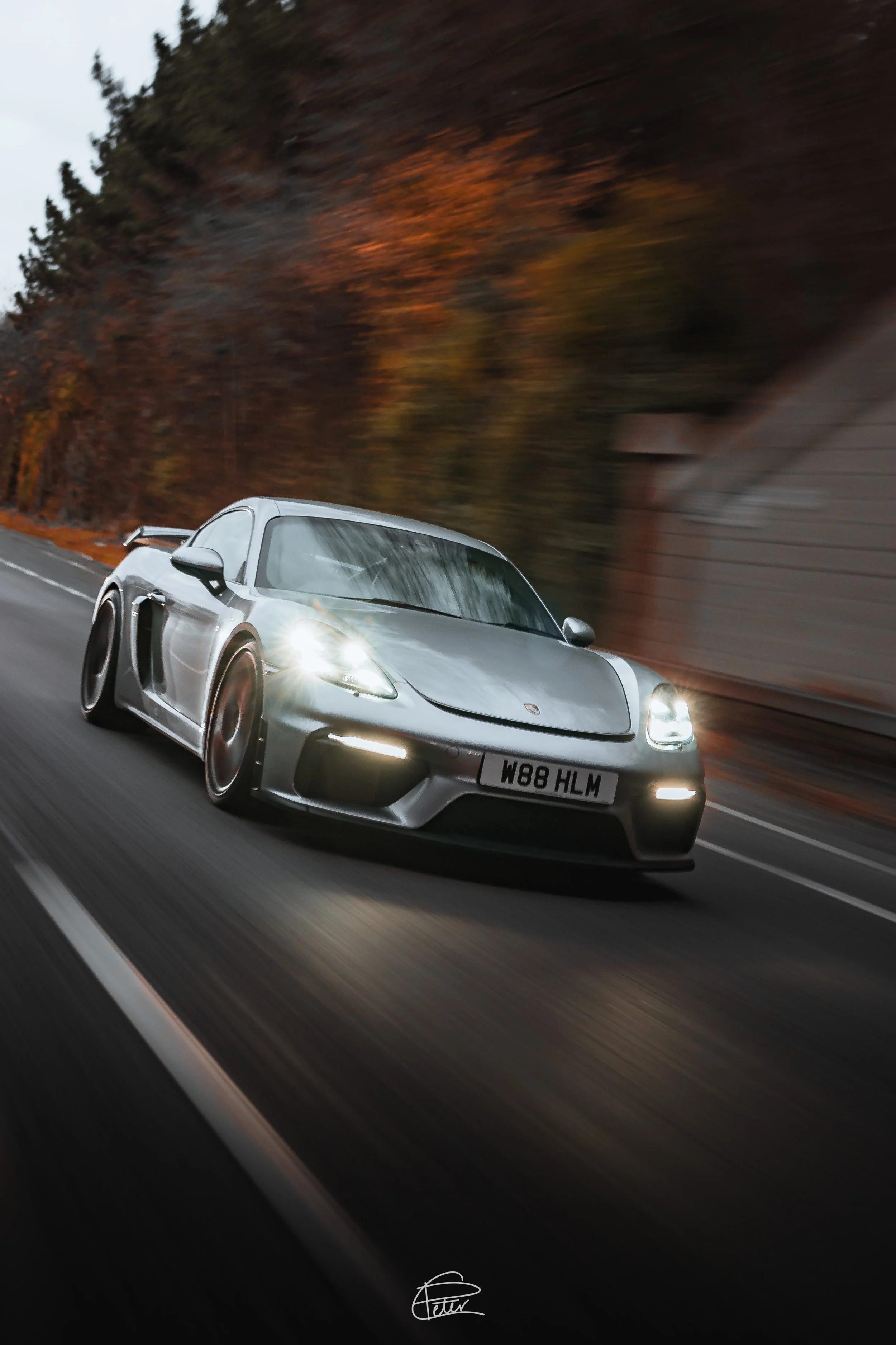 A silver sports car, resembling a Porsche, speeding down a highway surrounded by autumn trees with fall foliage, during daytime.