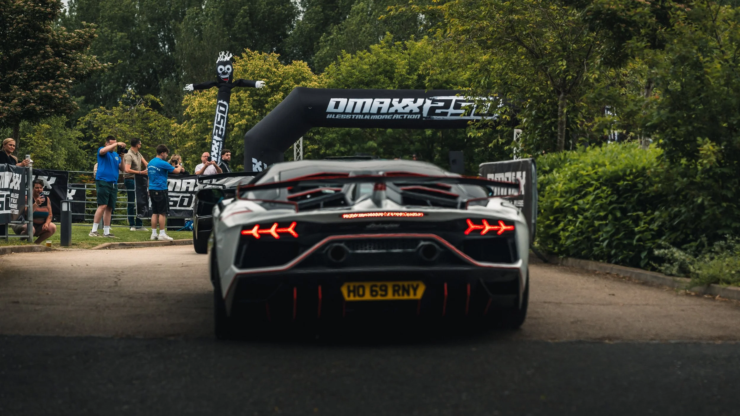 A white sports car with red accents and a visible license plate reading 'HO 69 RNY' driving on a road with green trees on either side. In the background, people are gathered near a black inflatable arch with white text and a black figure with a comic