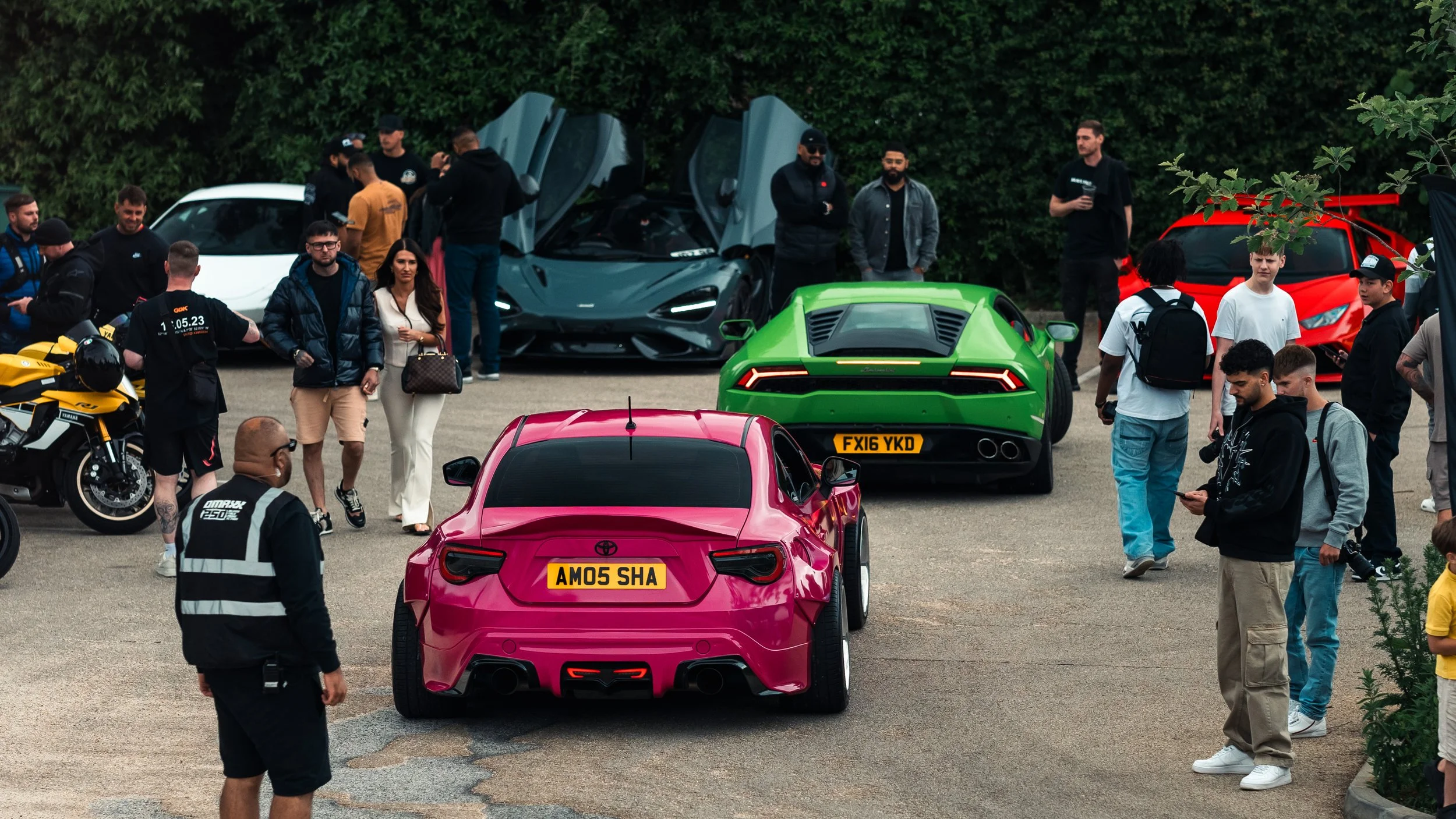 A group of people gathered around various luxury and sports cars at an outdoor car event, with some individuals taking photos and others conversing.