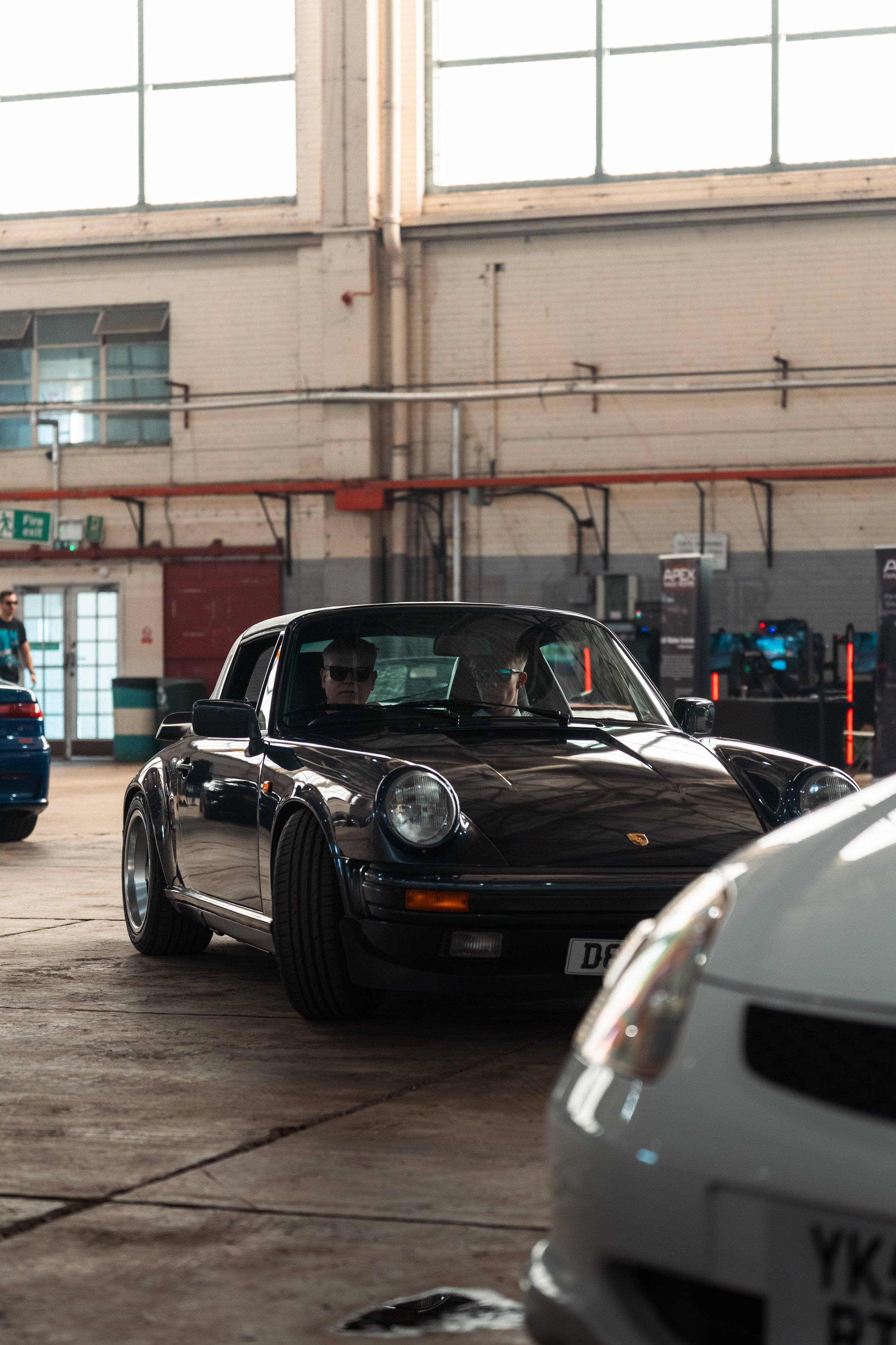 A black Porsche 911 convertible is parked inside an industrial building with large windows and exposed brick walls. Two people are sitting inside the car, wearing dark sunglasses.