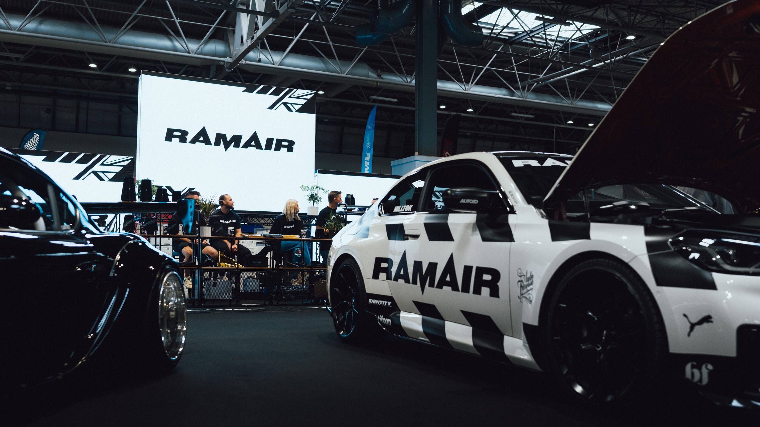 Multiple racing cars with 'RAM AIR' branding on display at an indoor auto show, with a large illuminated 'RAM AIR' sign and several people seated behind a viewing area.