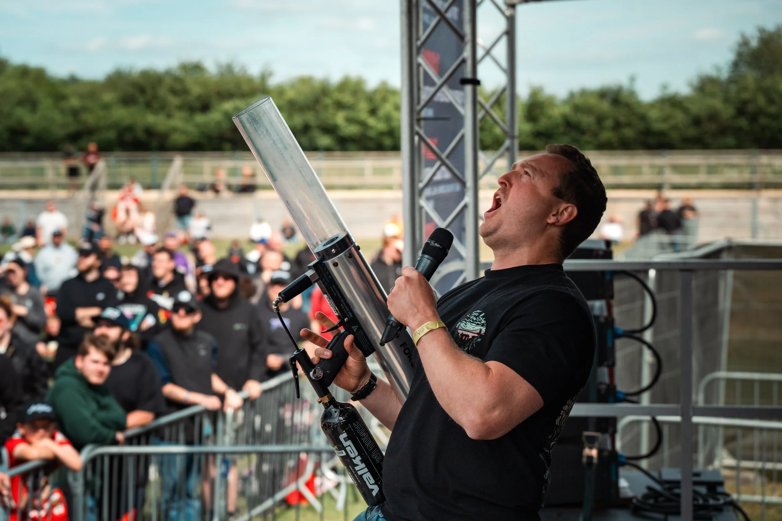 A man speaking into a microphone while holding a scooter at an outdoor event with an audience in the background.