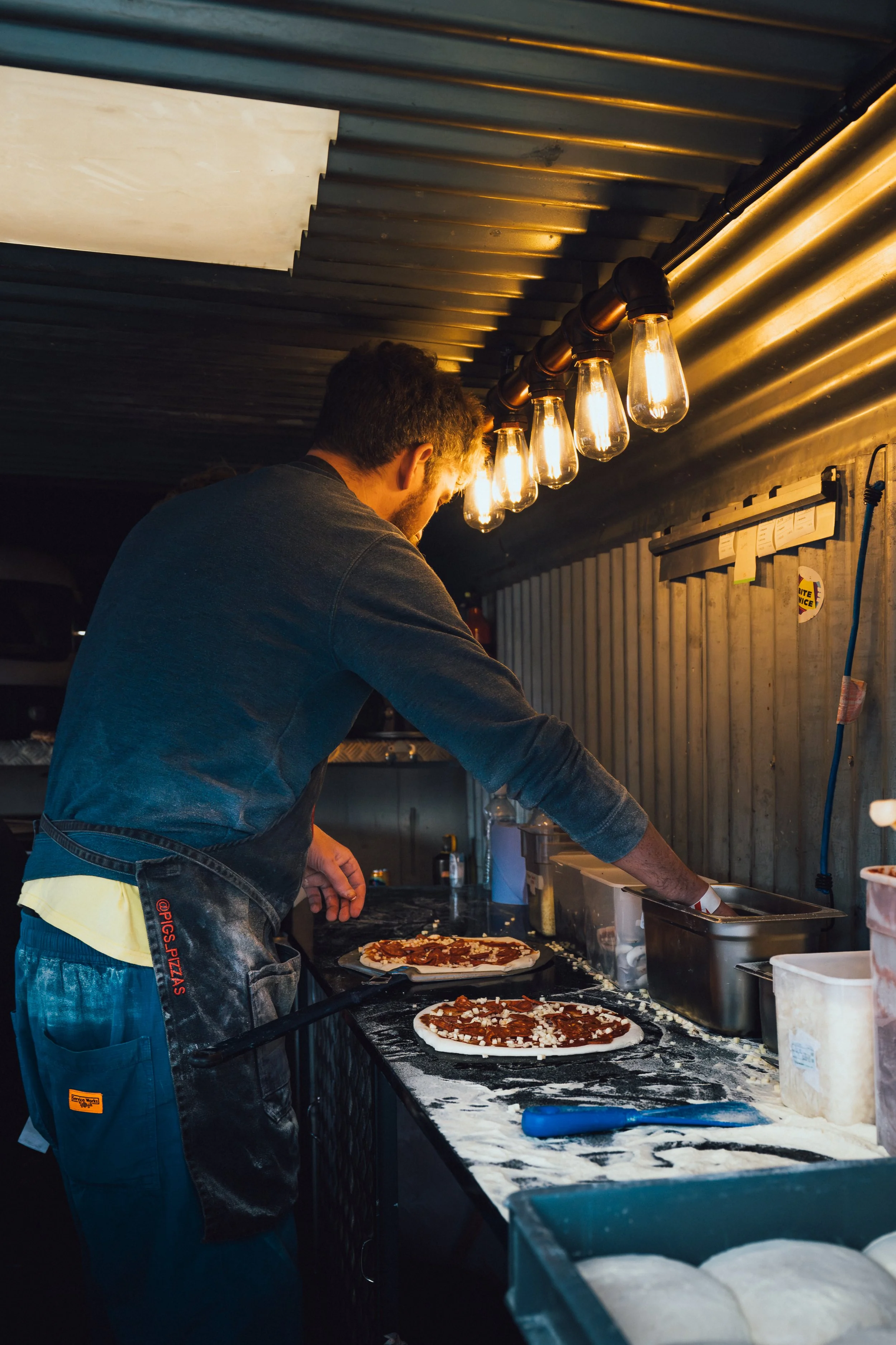 A person preparing pizzas in a kitchen with a corrugated metal wall and industrial-style lighting. The person is adding toppings to pre-made pizza bases on a black countertop, with other pizza bases and kitchen supplies around.