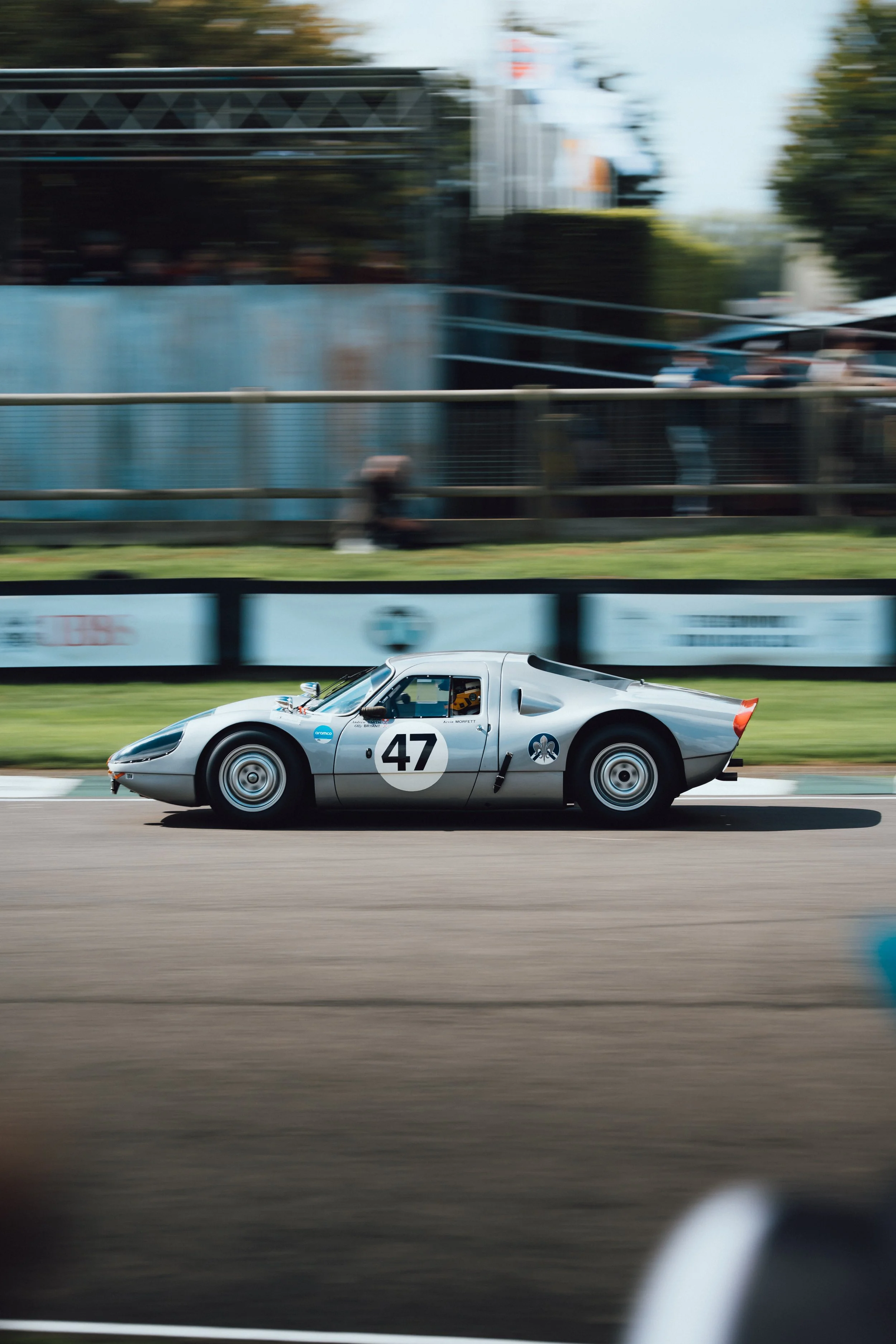 A silver race car with the number 47 on its side driving on a race track. The background shows a blurred view of spectators and structures, indicating motion and speed.