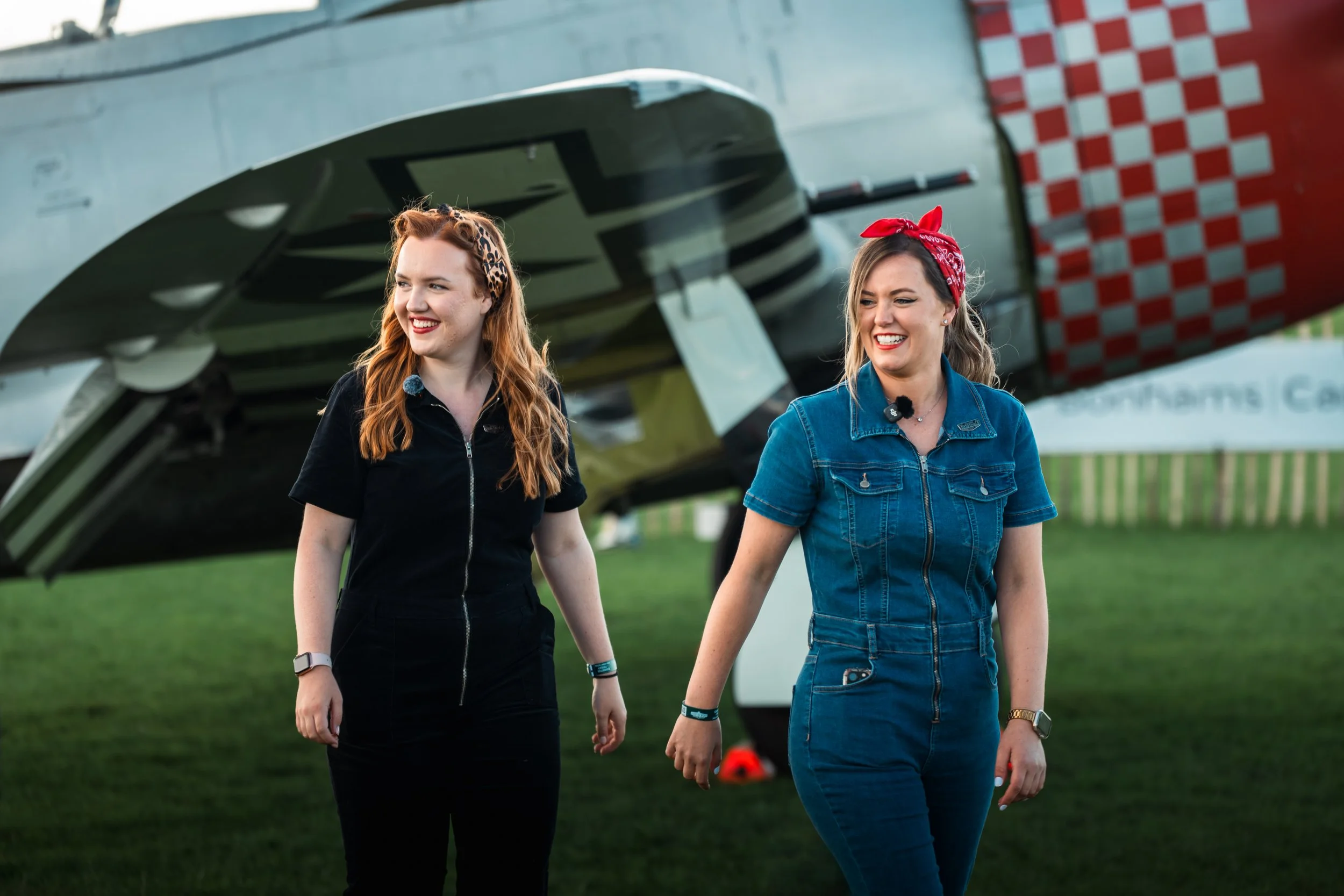 Two smiling women holding hands outdoors near a helicopter, wearing casual clothing and hair accessories, with a grassy area and a fence in the background.