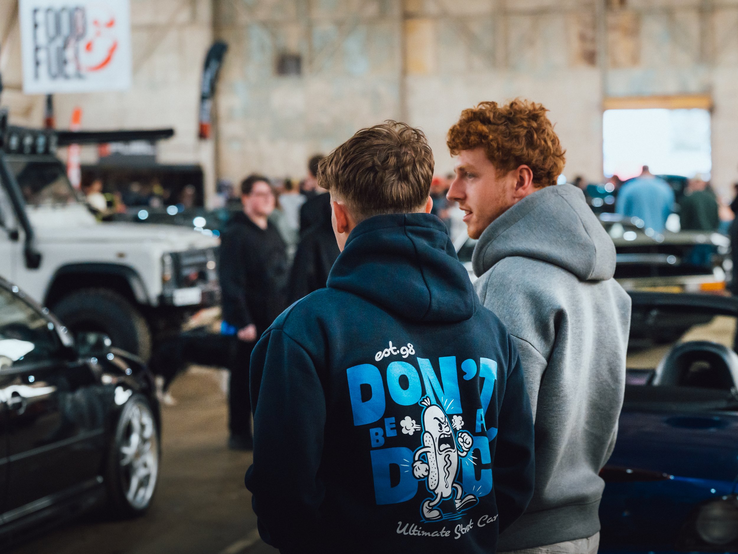 Two young men with curly hair are having a conversation in an indoor car exhibition or auto show, surrounded by vintage and modern cars, with a crowd in the background.
