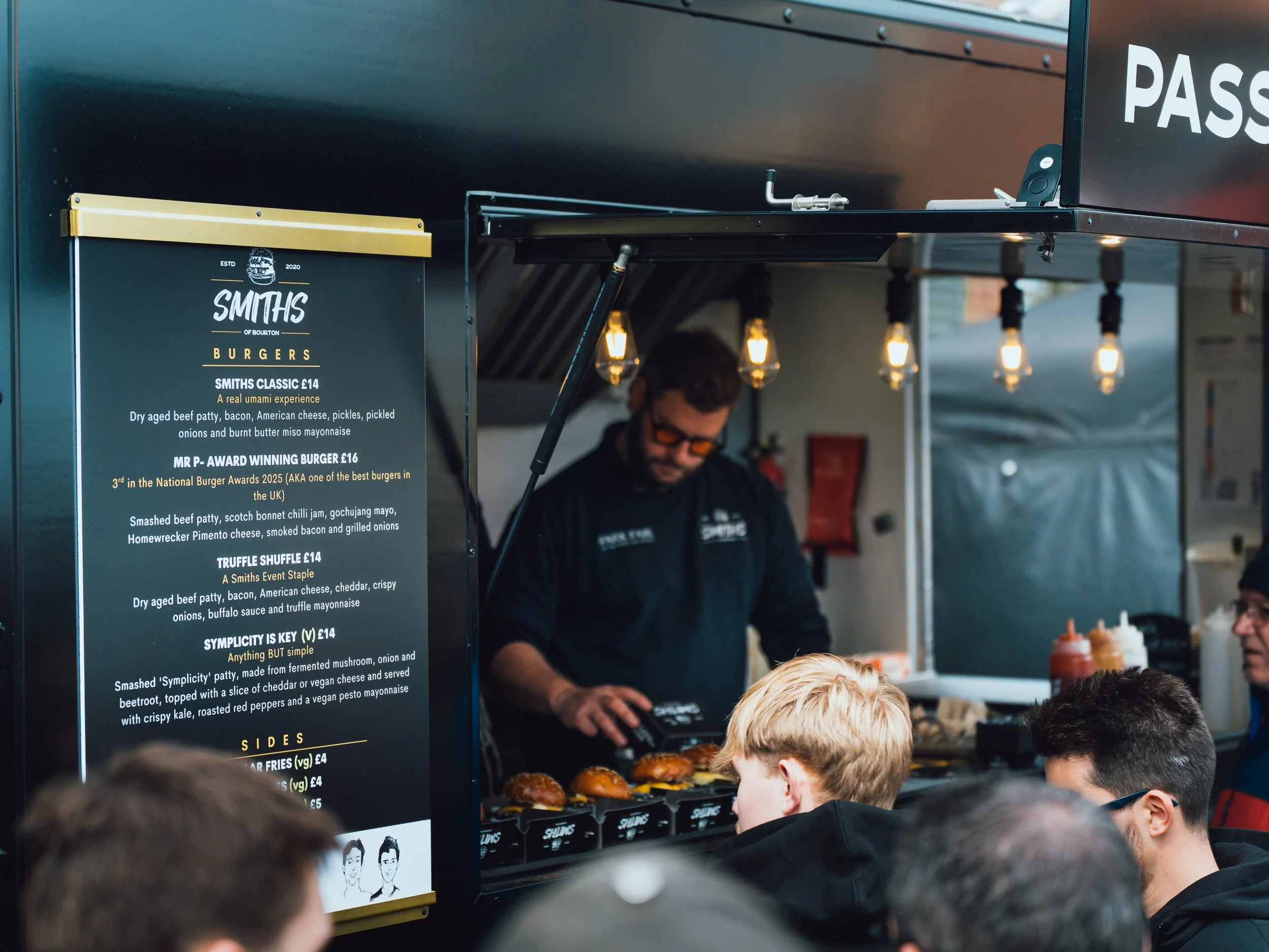 A food stand menu with multiple burger options and sides, and people in line in front of it, with the stand's staff preparing food behind the counter.