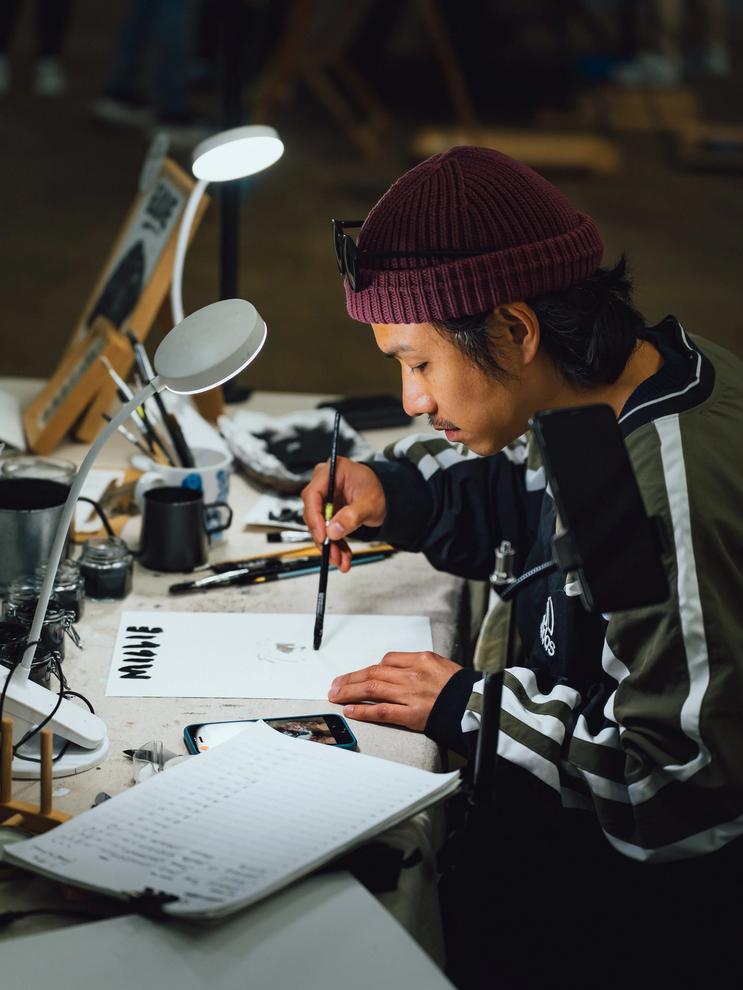 A person wearing a maroon beanie and black sunglasses is sitting at a cluttered desk, writing in a notebook with various objects including a book, smartphone, and desk lamps around.