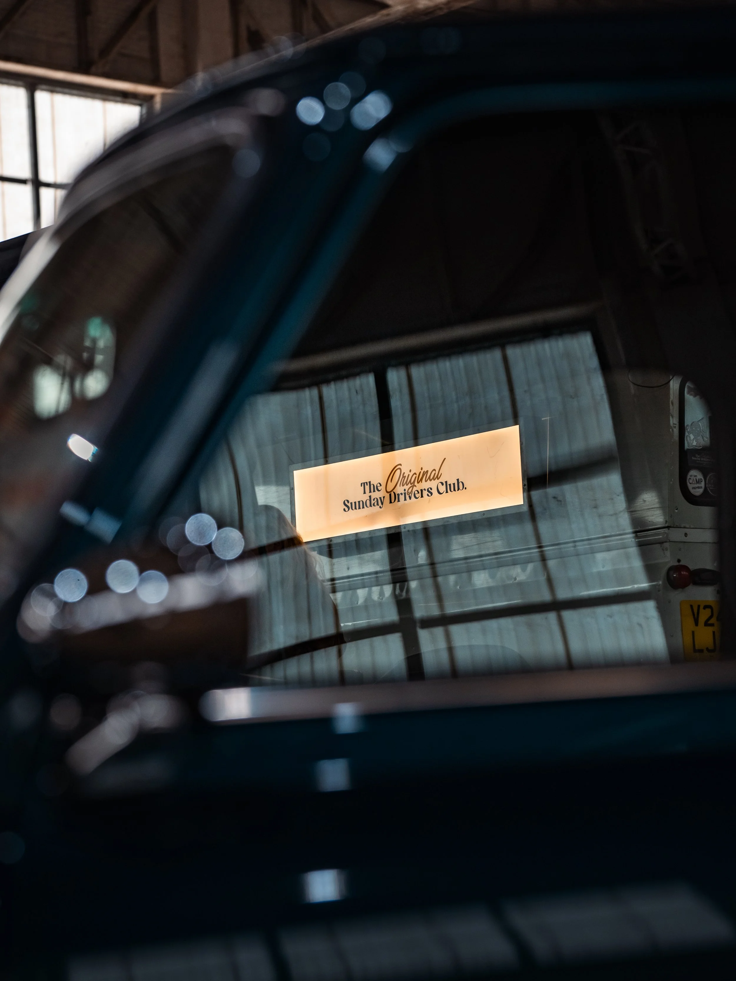 Photo showcasing the rear window of a vehicle displaying a sign that reads 'The Original Sunday Drivers Club,' with reflections and a wooden ceiling visible in the background.