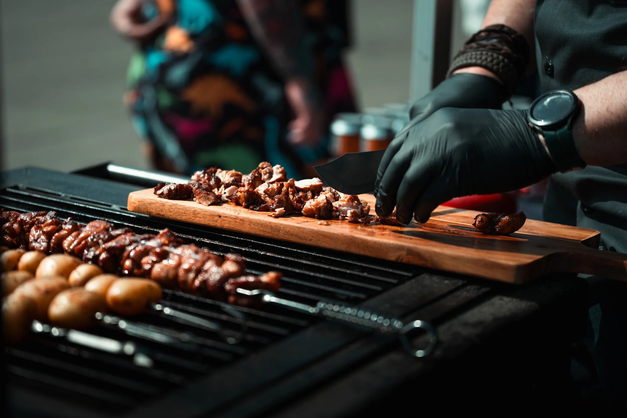 Person wearing black gloves slicing cooked meat on a wooden cutting board at a barbecue grill station.