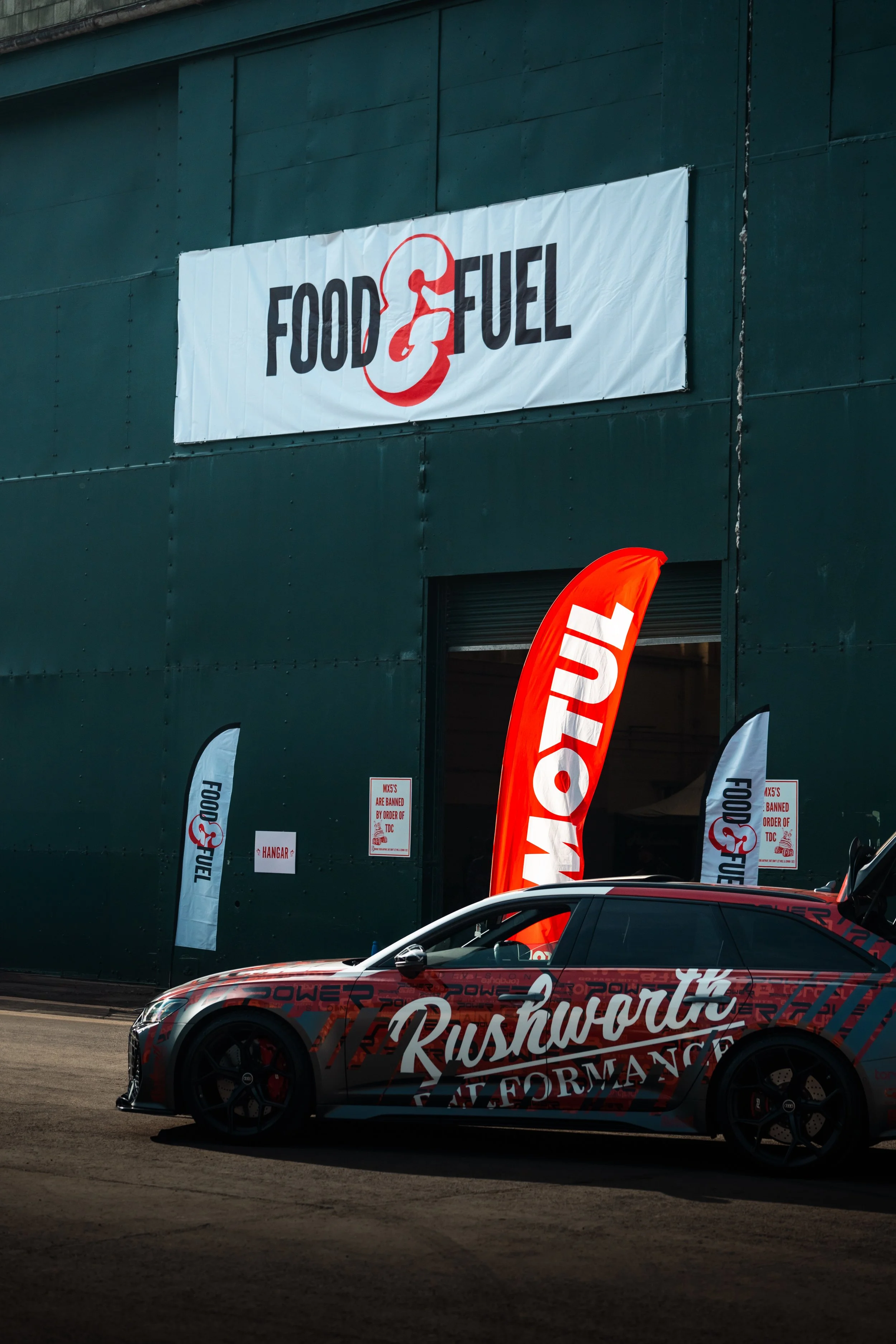 A racing car with the words "Rushworth Performance" on the side, parked in front of a large sign that says "Food & Fuel". The scene is set outside a building with flags and banners promoting the event.