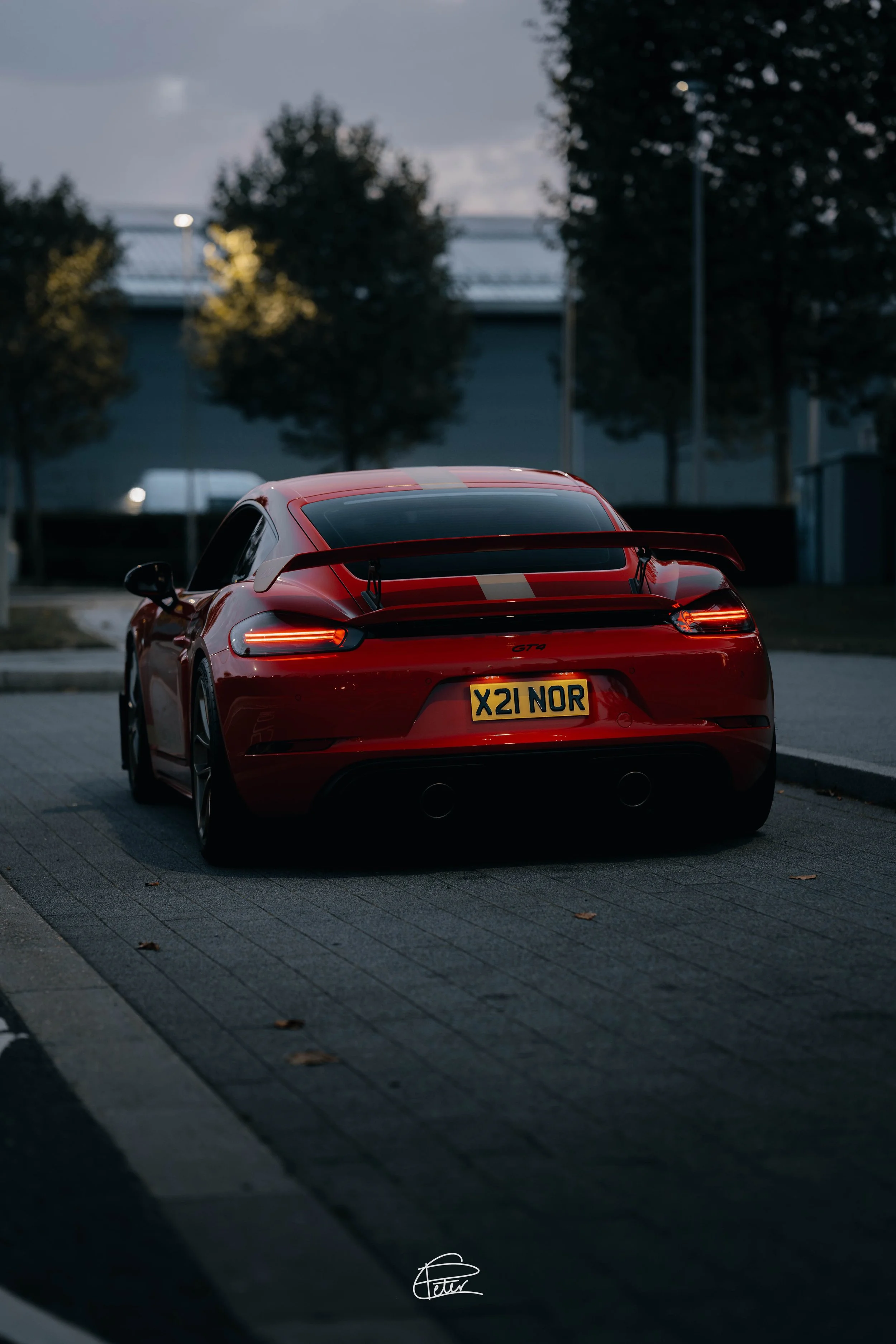 A red sports car parked on a paved area during dusk, with trees and a building in the background.