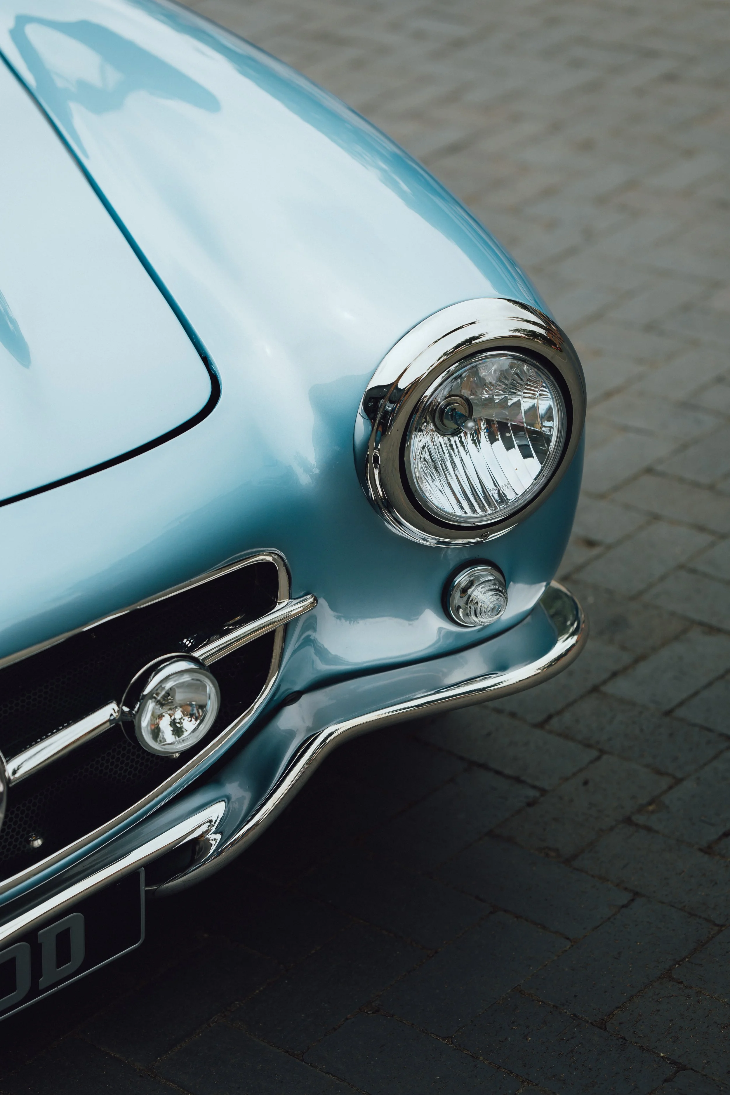 Close-up of the front part of a light blue vintage car, showing the headlight, grille, and part of the bumper, parked on a brick pavement.