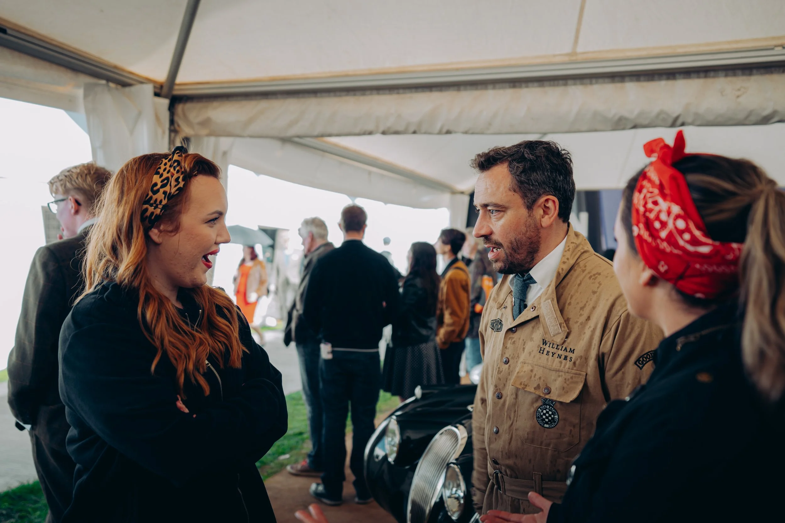 A woman with red hair and a leopard print headband talking to a man in a beige uniform at an outdoor event under a tent, with other people in the background.