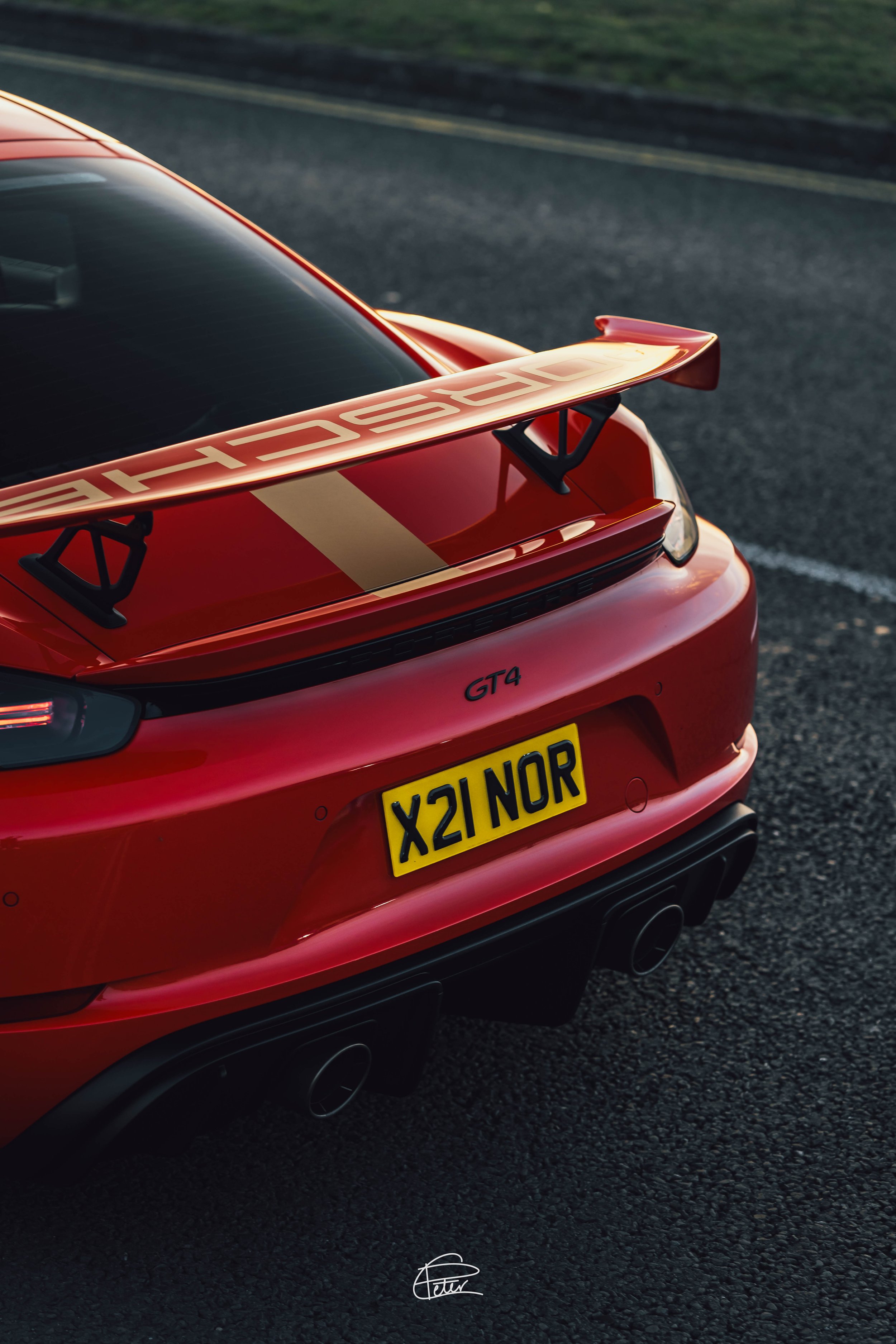 Close-up of the rear of a red Porsche GT4 car with a large rear wing, dual exhaust pipes, and a license plate reading 'X21 NOR', parked on a dark gravel surface.