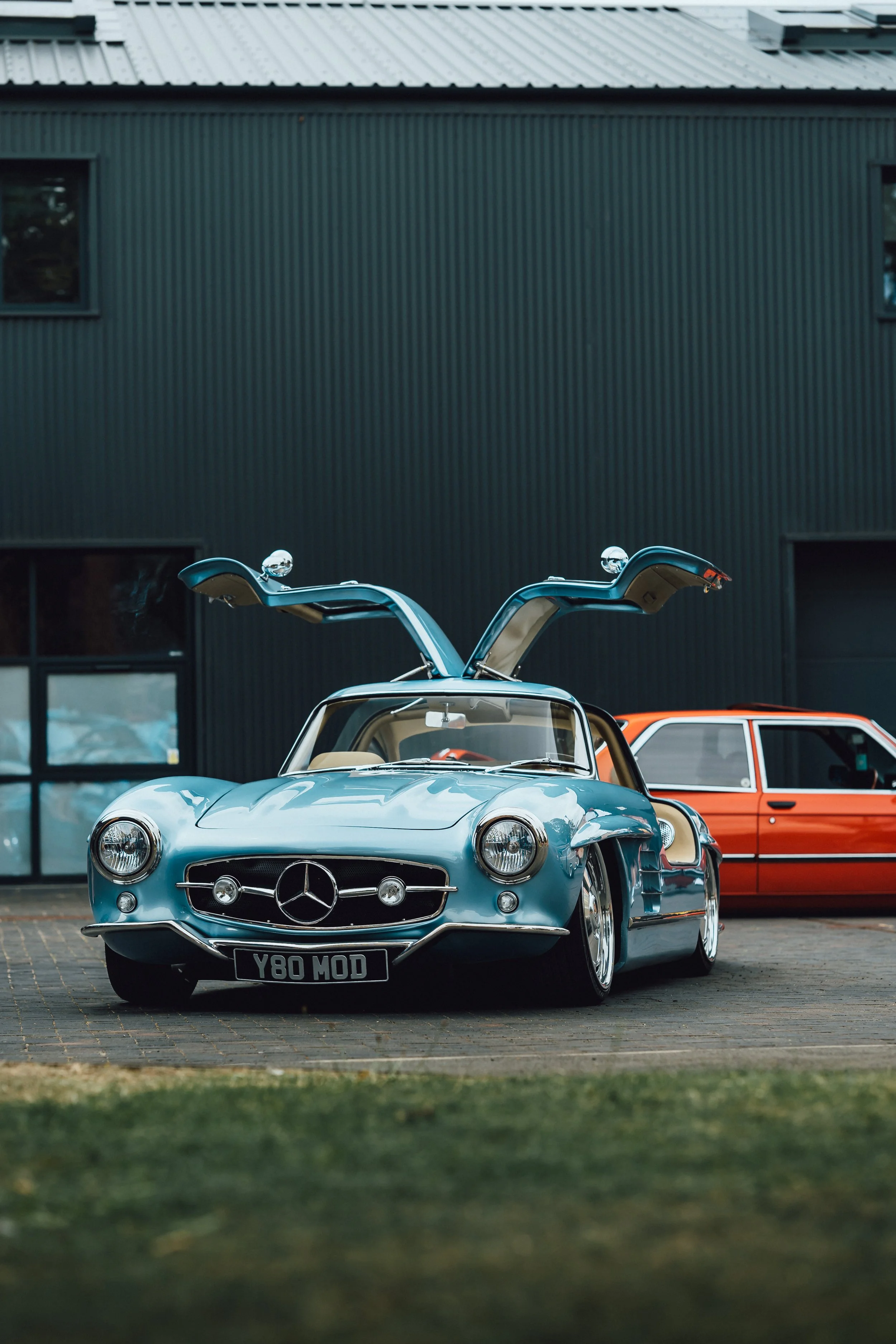 A vintage silver Mercedes-Benz sports car with gullwing doors open, parked in front of a modern dark building, with a red classic car beside it.