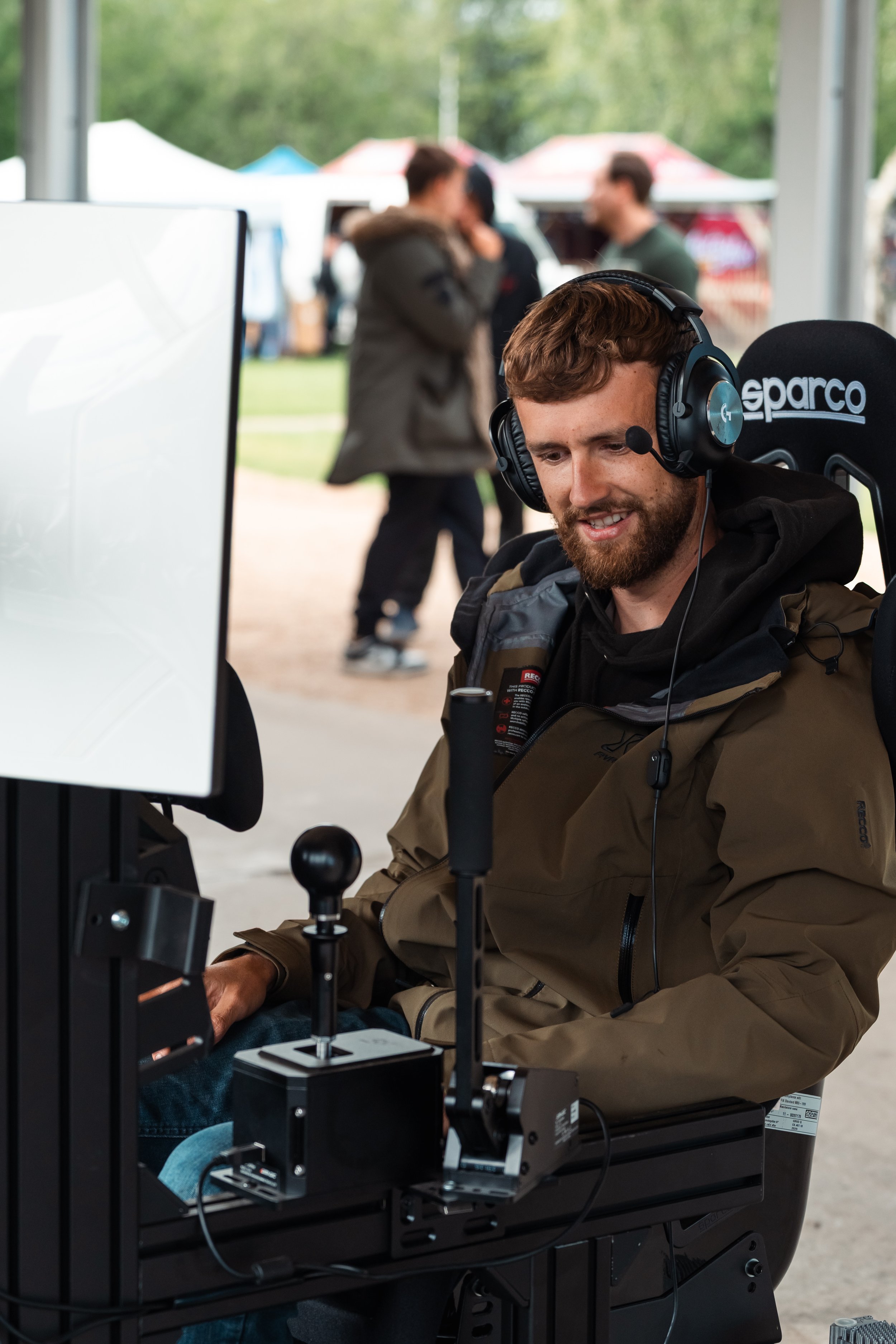 A man with a beard and brown hair wearing a headset, sitting at a control station with various equipment, smiling and working. In the background, a group of blurred people are outdoors near tents.