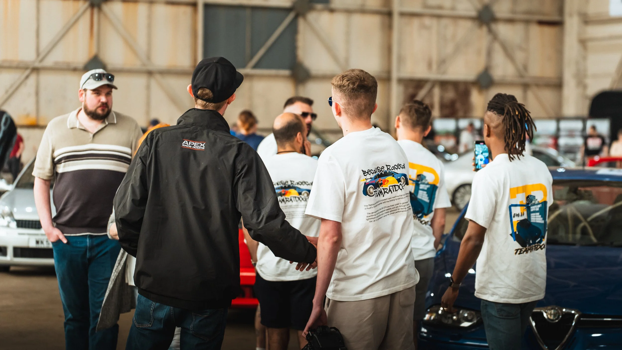 A group of people are gathered indoors at a car event, some wearing white T-shirts with colorful race car graphics, while others are in casual clothing. They appear to be talking, taking photos, and observing cars in the background.