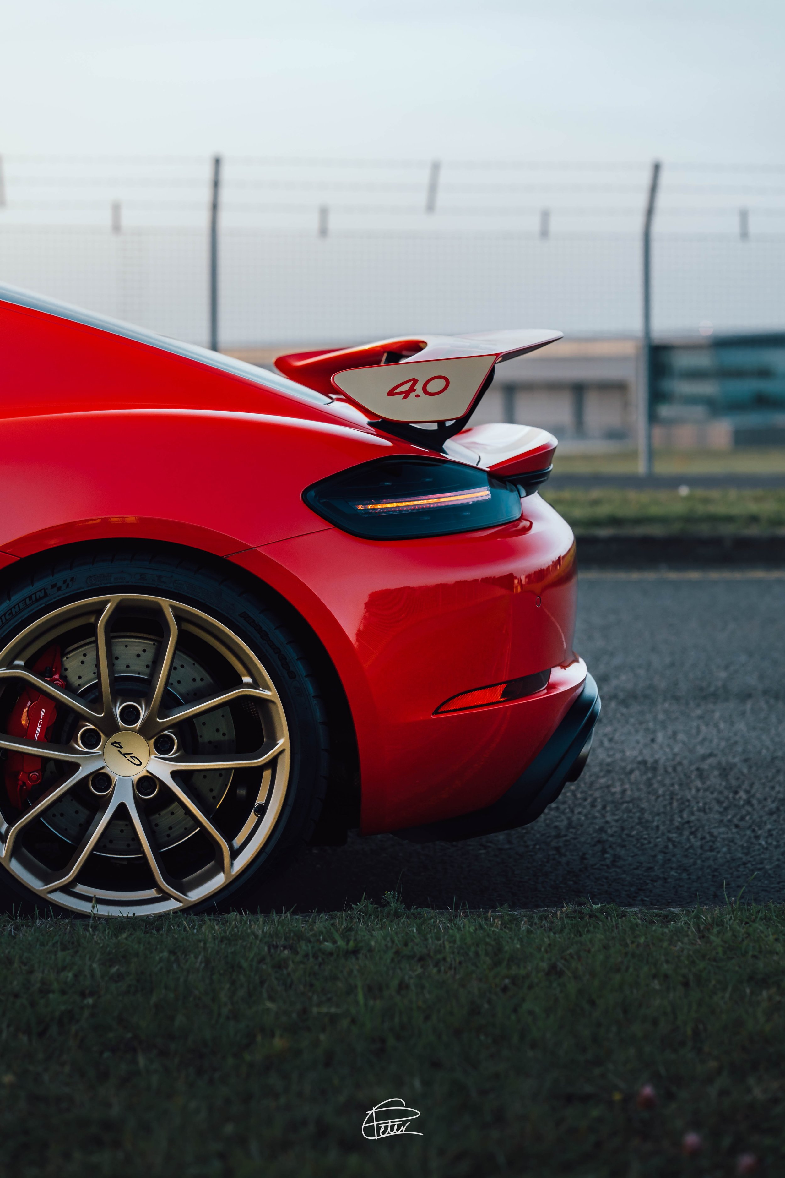 Close-up of the rear part of a red sports car, showing a gold alloy wheel, a black brake caliper, and a rear wing with '4.0' written on it, parking on a grassy area near a racetrack with fencing in the background.