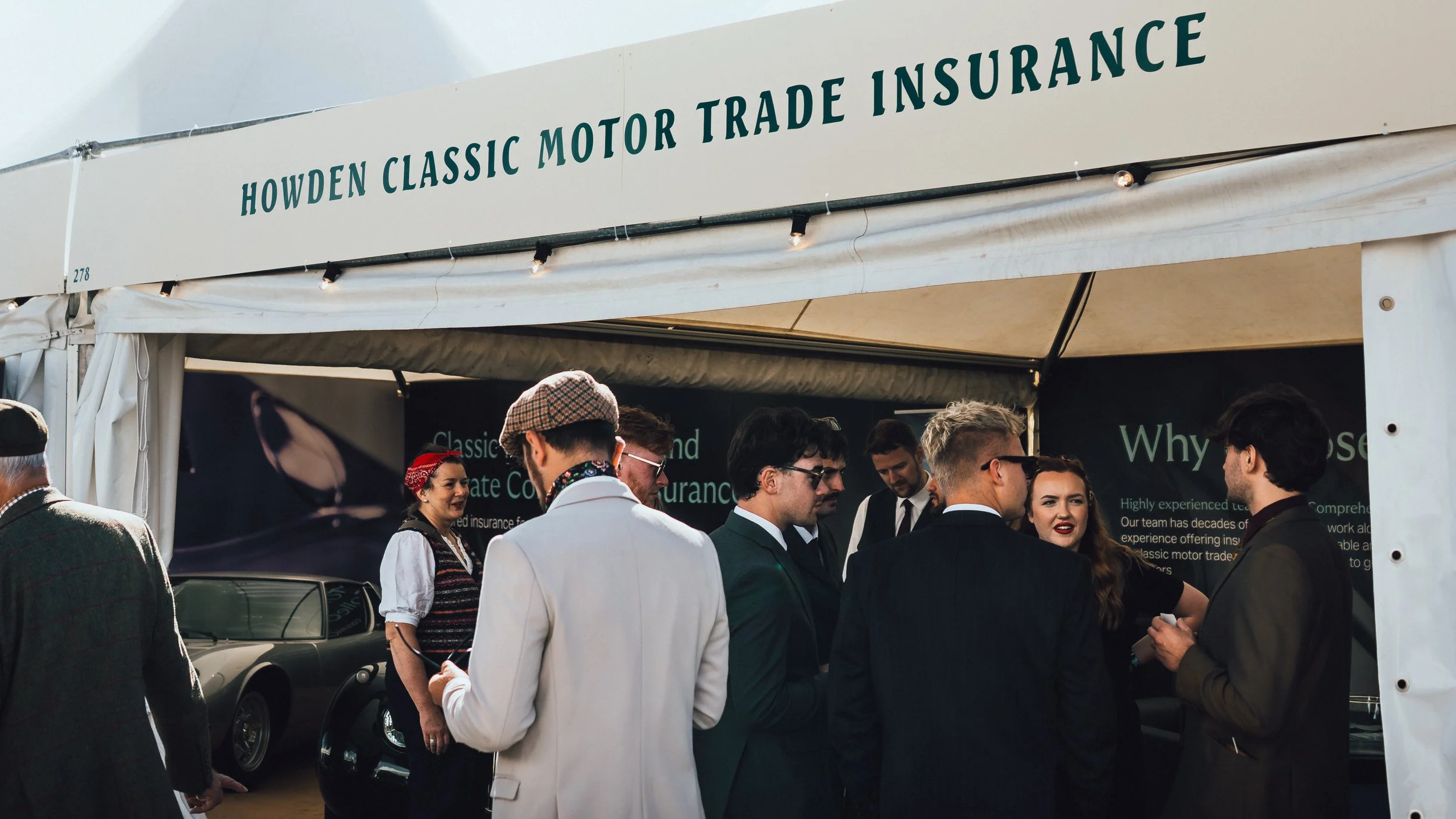 People gathered under a white tent at the Howden Classic Motor Trade Insurance event, engaging in conversations, with a car partially visible on the left side.