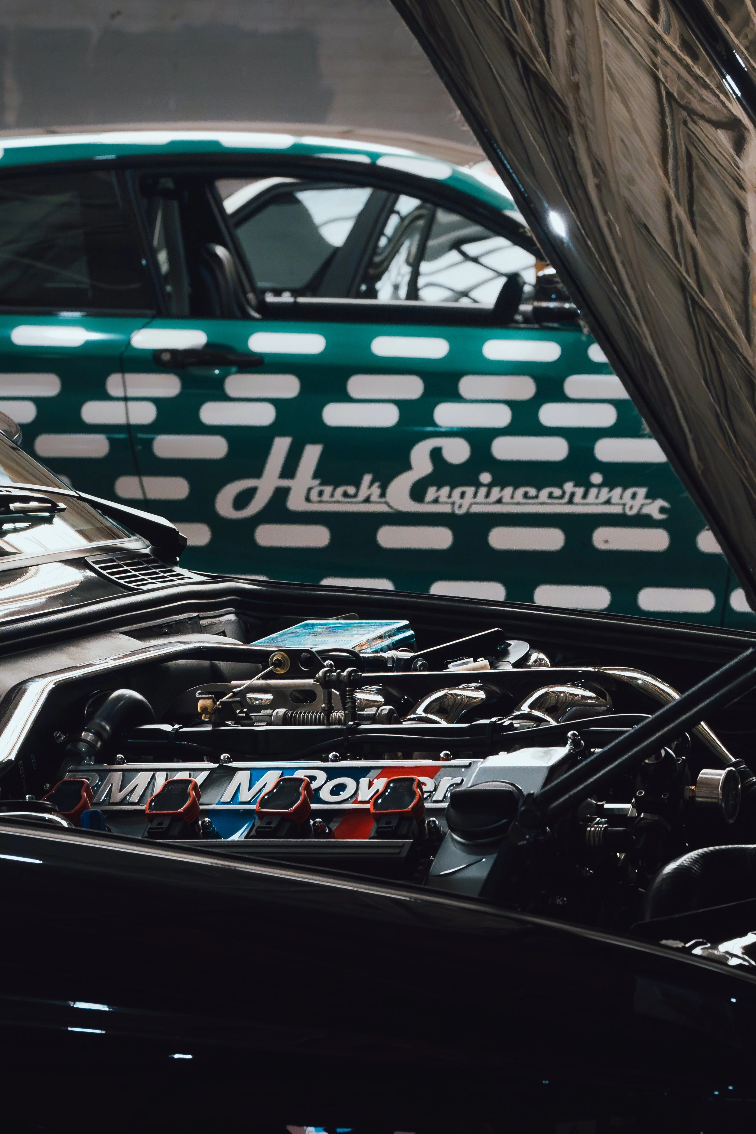 The image shows the engines of two race cars with the hoods open, revealing their detailed mechanical parts and branding.