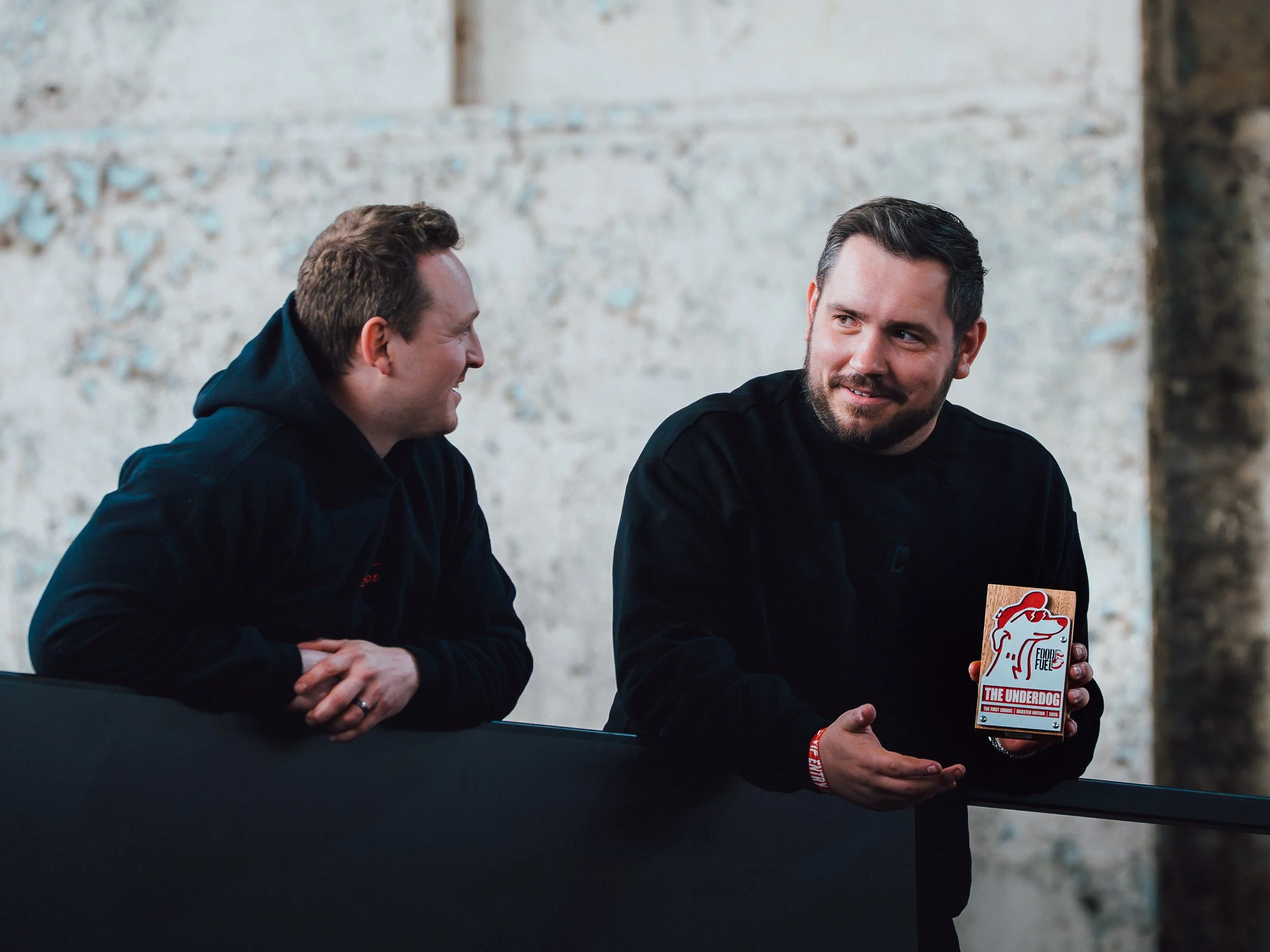Two men sitting at a table, engaged in conversation, with one holding a sign that says 'The Underdog' and features a logo of a dog's head. The setting appears to be indoors with a rustic, textured wall in the background.