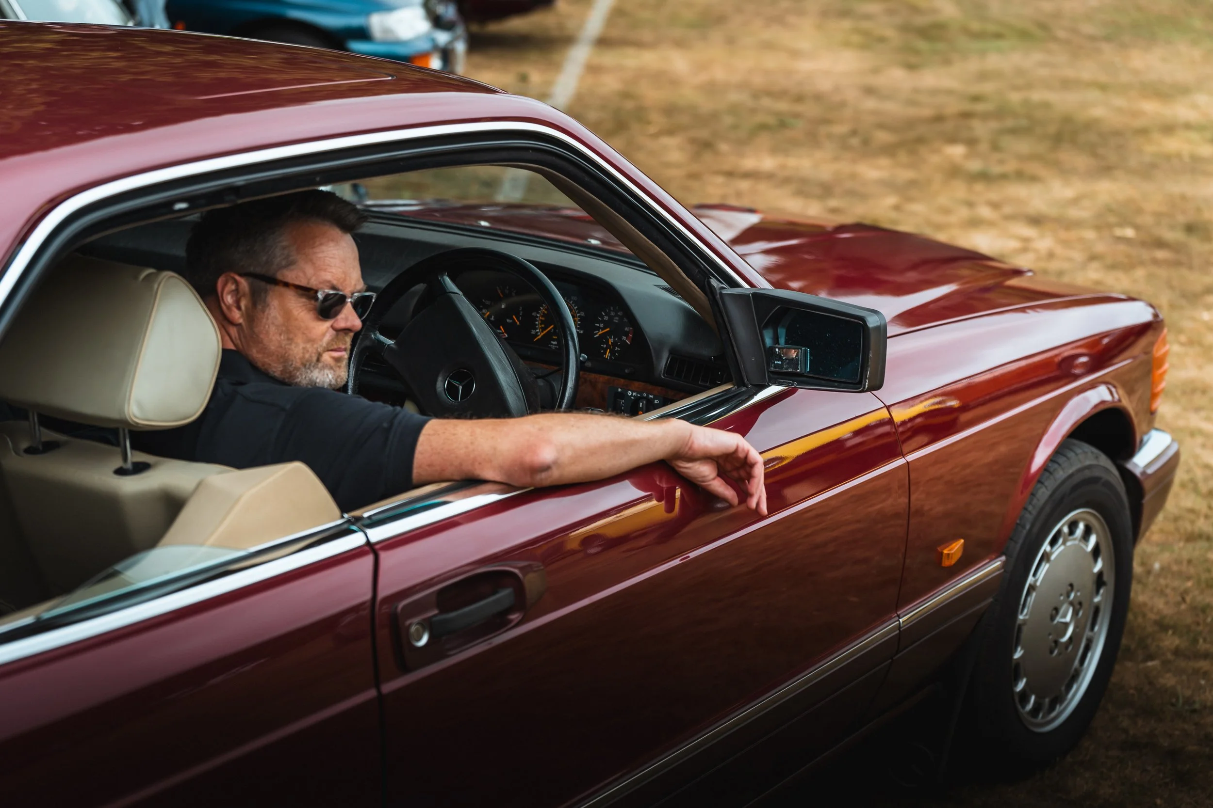A man with sunglasses sitting in the driver's seat of a vintage red Mercedes-Benz car, leaning out of the window with a serious expression on his face. The car is parked outdoors on a grassy area.