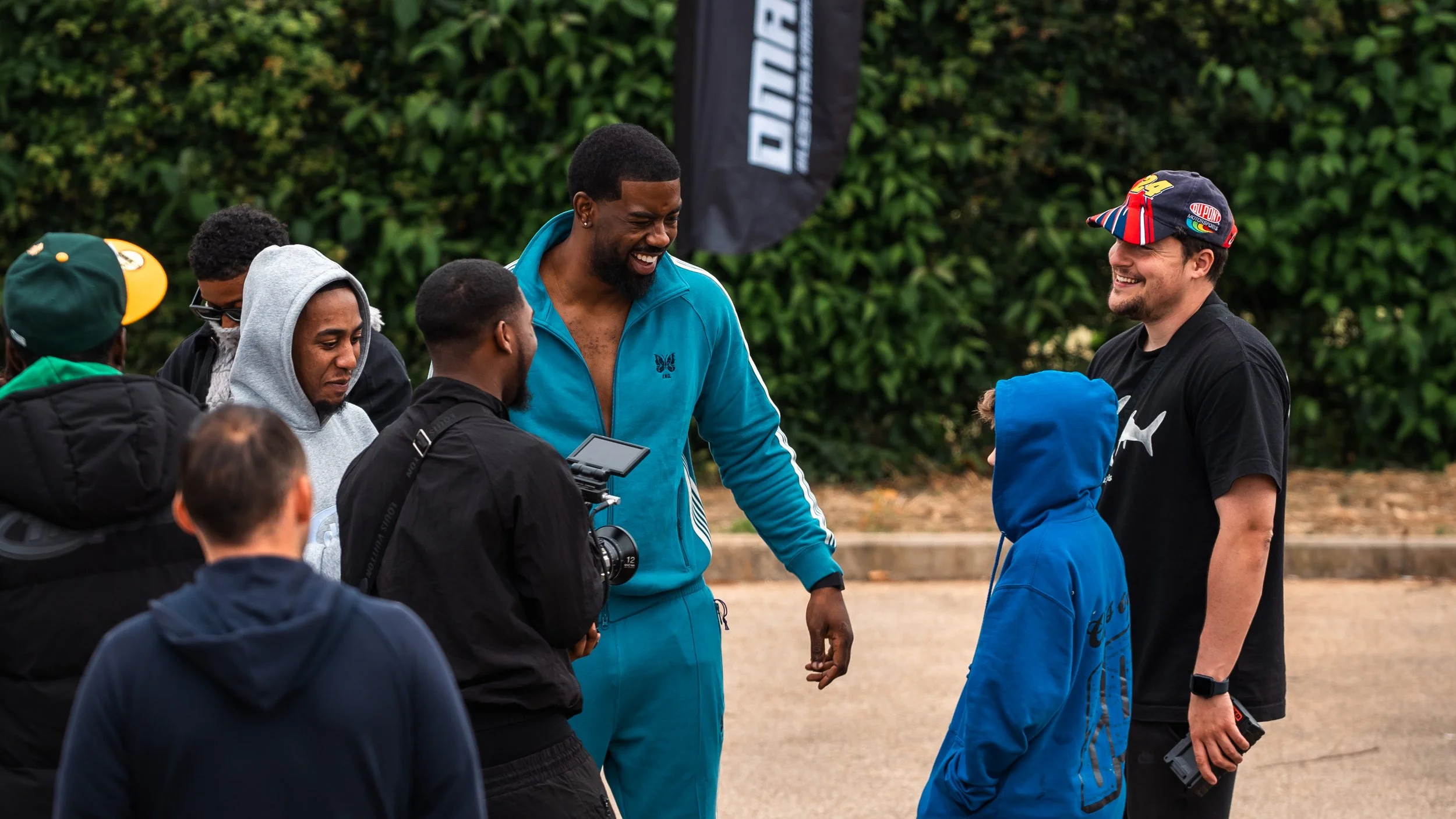 Group of young people and an adult man gathering outside, engaging in conversation and smiling. The setting appears to be a casual outdoor event with green foliage in the background.