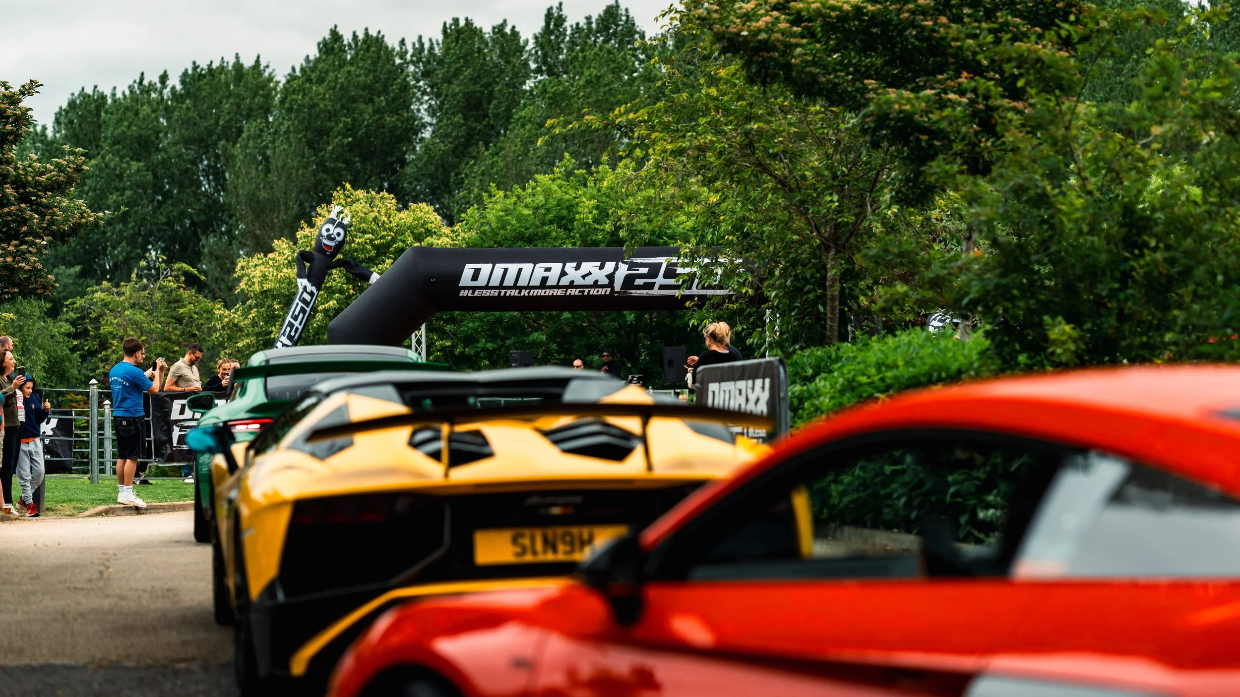 Multiple luxury sports cars parked outdoors at a car event, with a crowd taking photos. In the background, a person in a mascot costume stands near a large black inflatable arch with the text 'OMAXX 25' and 'A BLESS TALK MORE ACTION'.