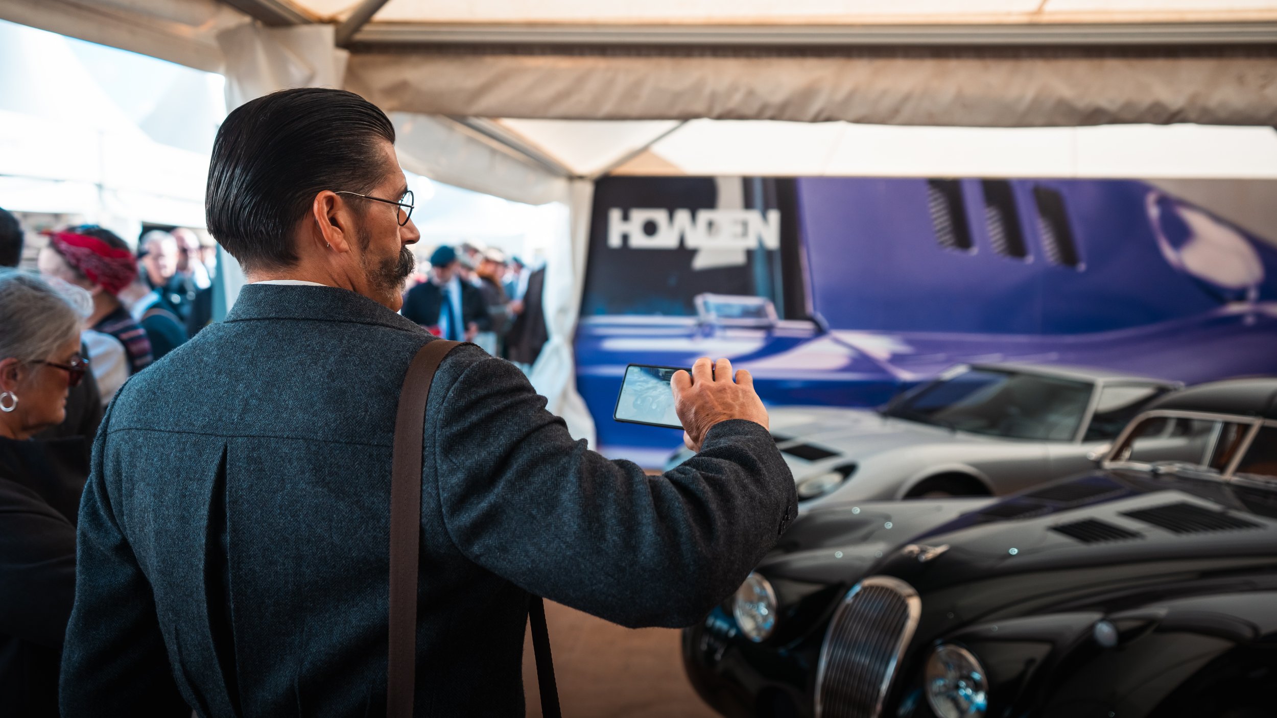 A man taking a photo of vintage cars at an outdoor event, with a crowd in the background and tents overhead.