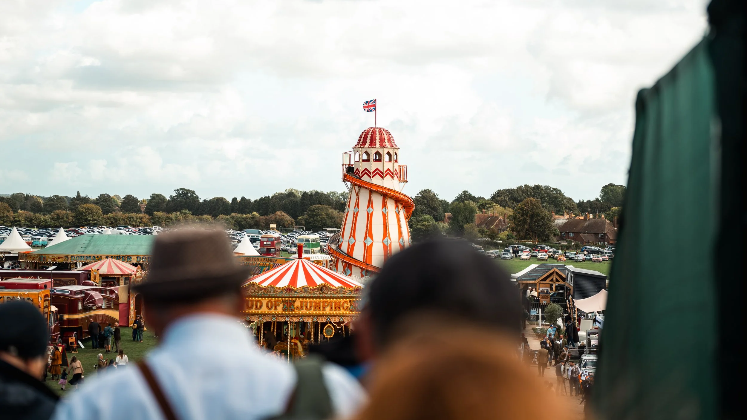 A carnival with a red and white striped up-and-down ride in the center, topped with a Union Jack flag. The carnival spills over a field with tents, stalls, and a parking lot filled with cars, under a cloudy sky.