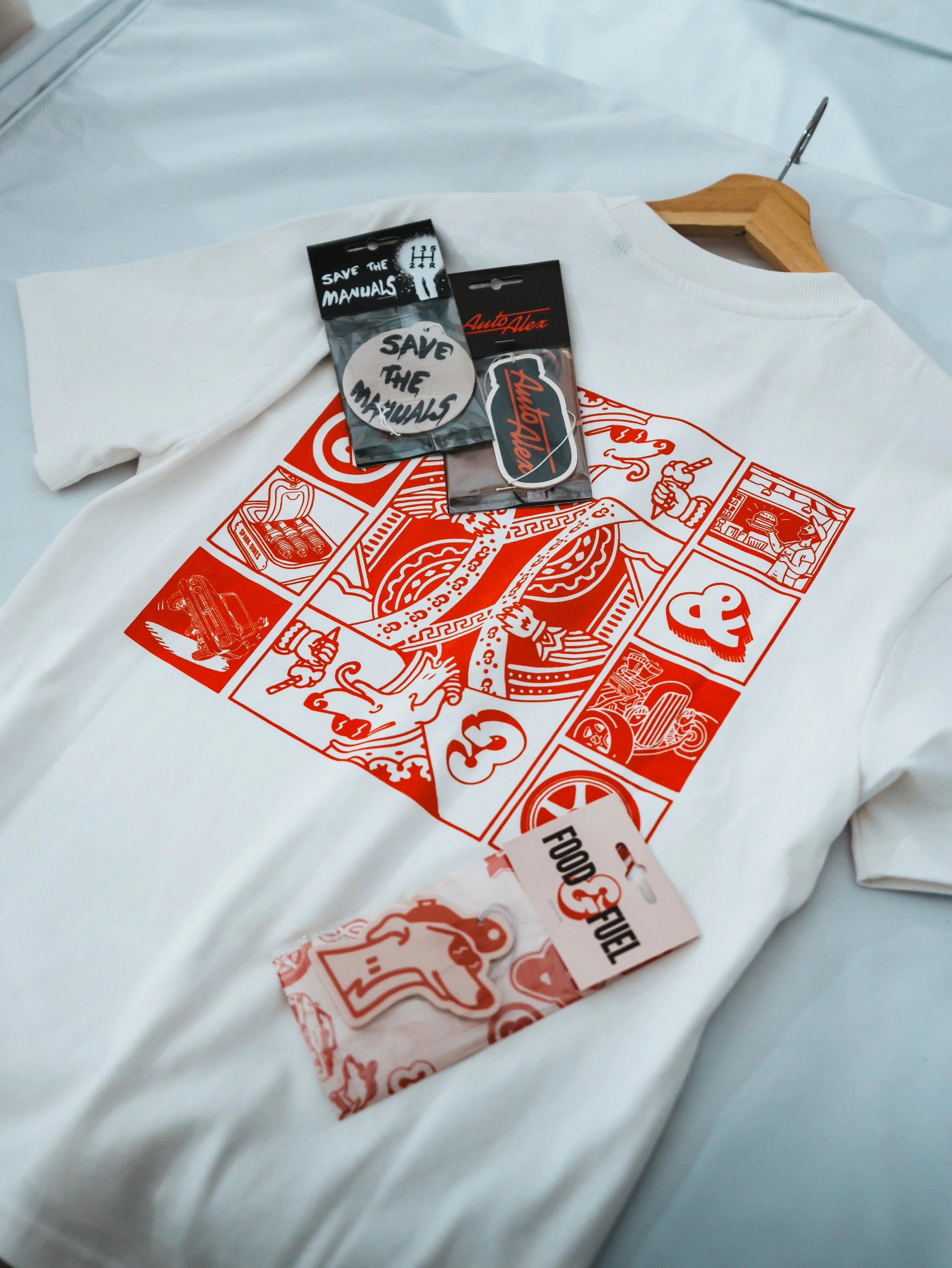 White T-shirt with red graphic print and two pins attached, placed on a hanger on a white surface.