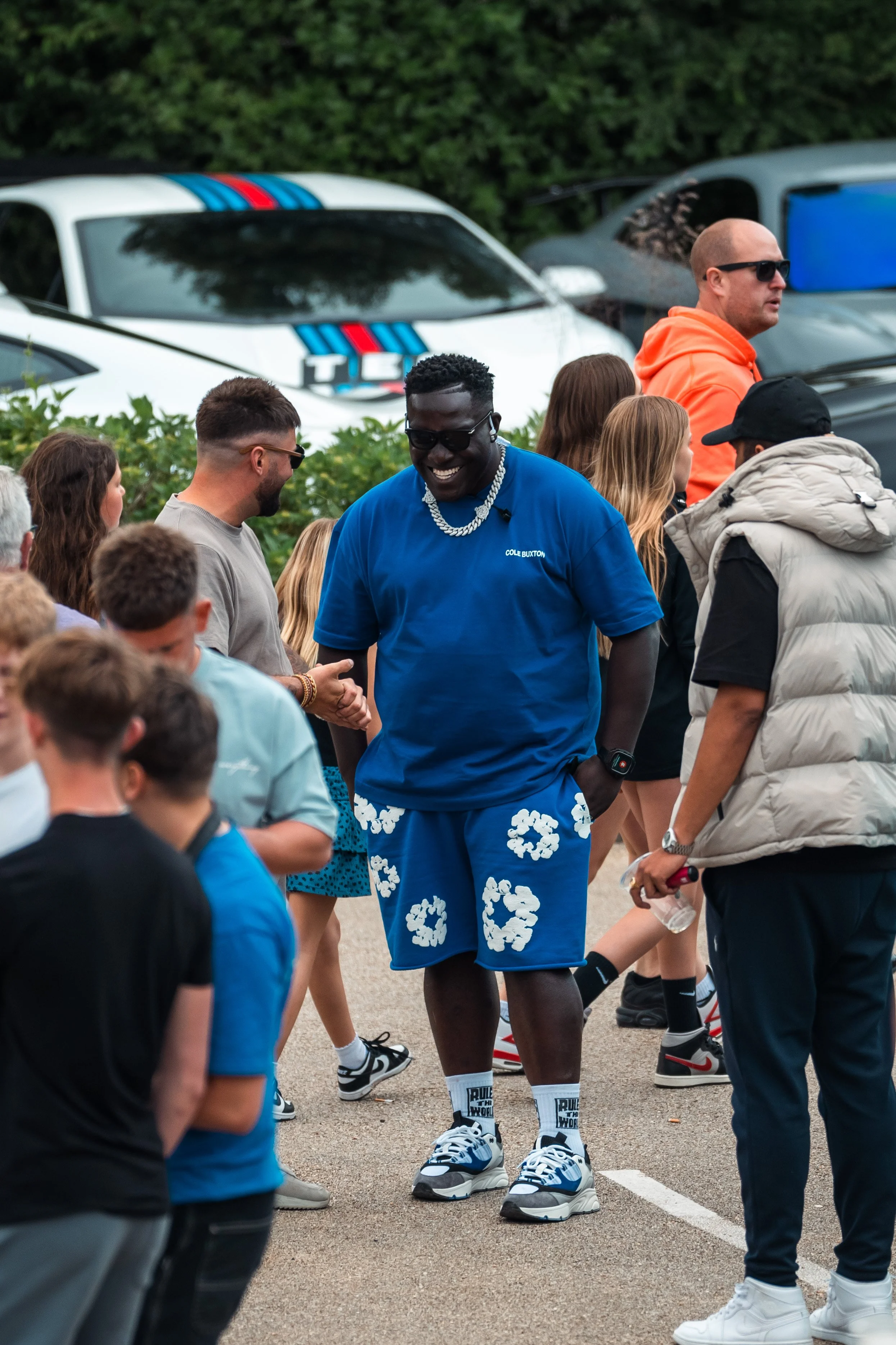 A group of people gathered outdoors, with a man in the center smiling, wearing a blue t-shirt with white floral patterns on the shorts, sunglasses, a chain necklace, and sneakers. Behind him are parked vehicles and a hedge, with some people engaged i