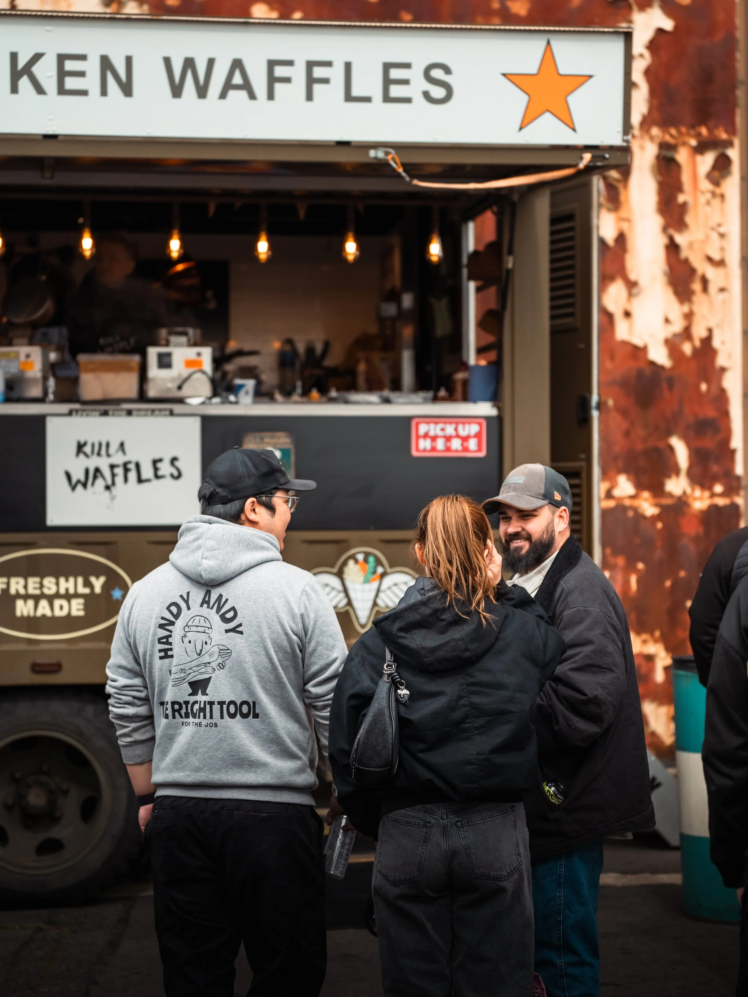 Group of people standing in front of a food truck with a sign that says "Ken Waffles" and a logo of an orange star. The food truck has a menu with waffle options, and there are three people visible in the foreground, engaged in conversation.