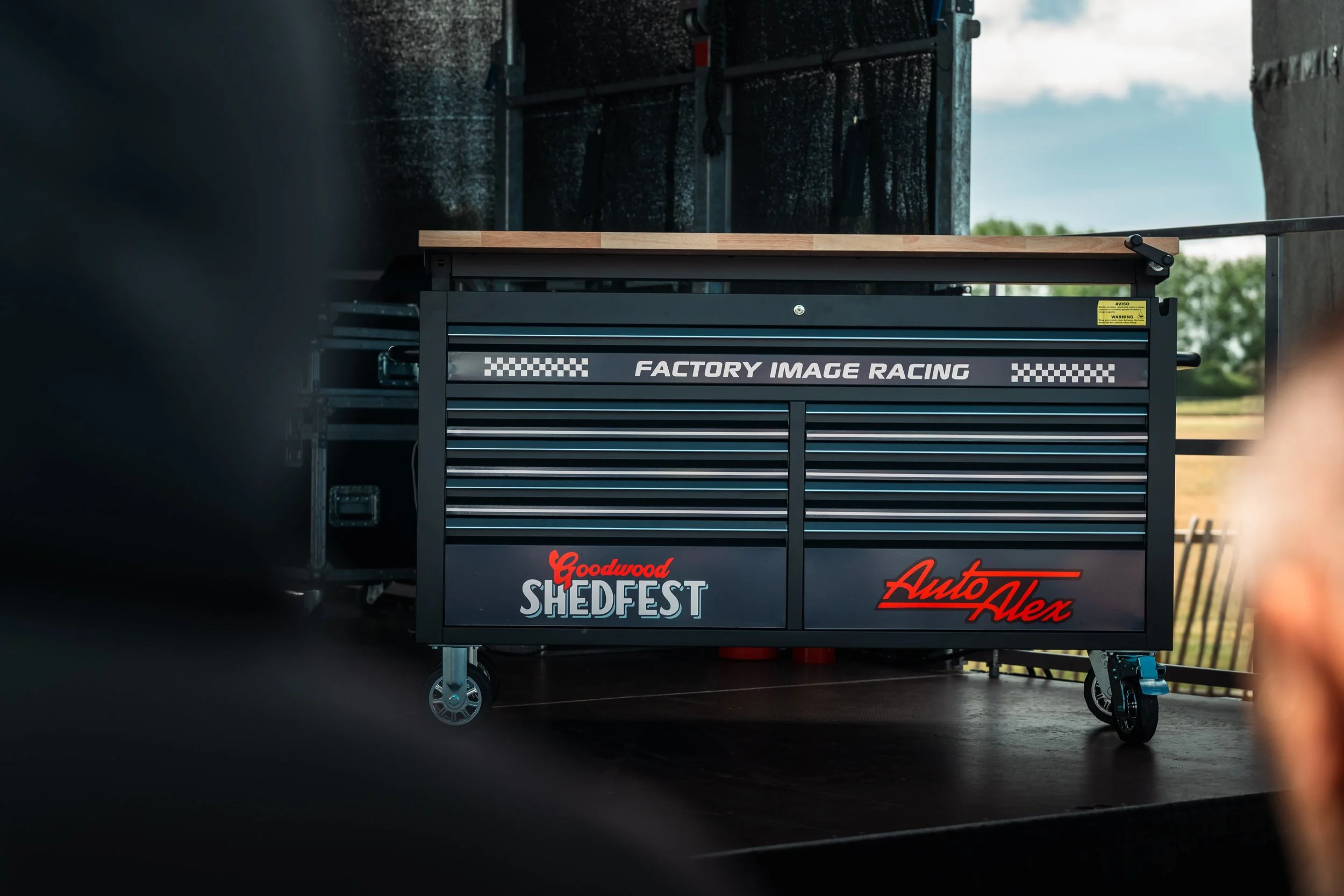 A tool chest with the logos of Goodwood Shedfest and AutoVex, situated outdoors on a concrete surface, with a background of sky and greenery visible.